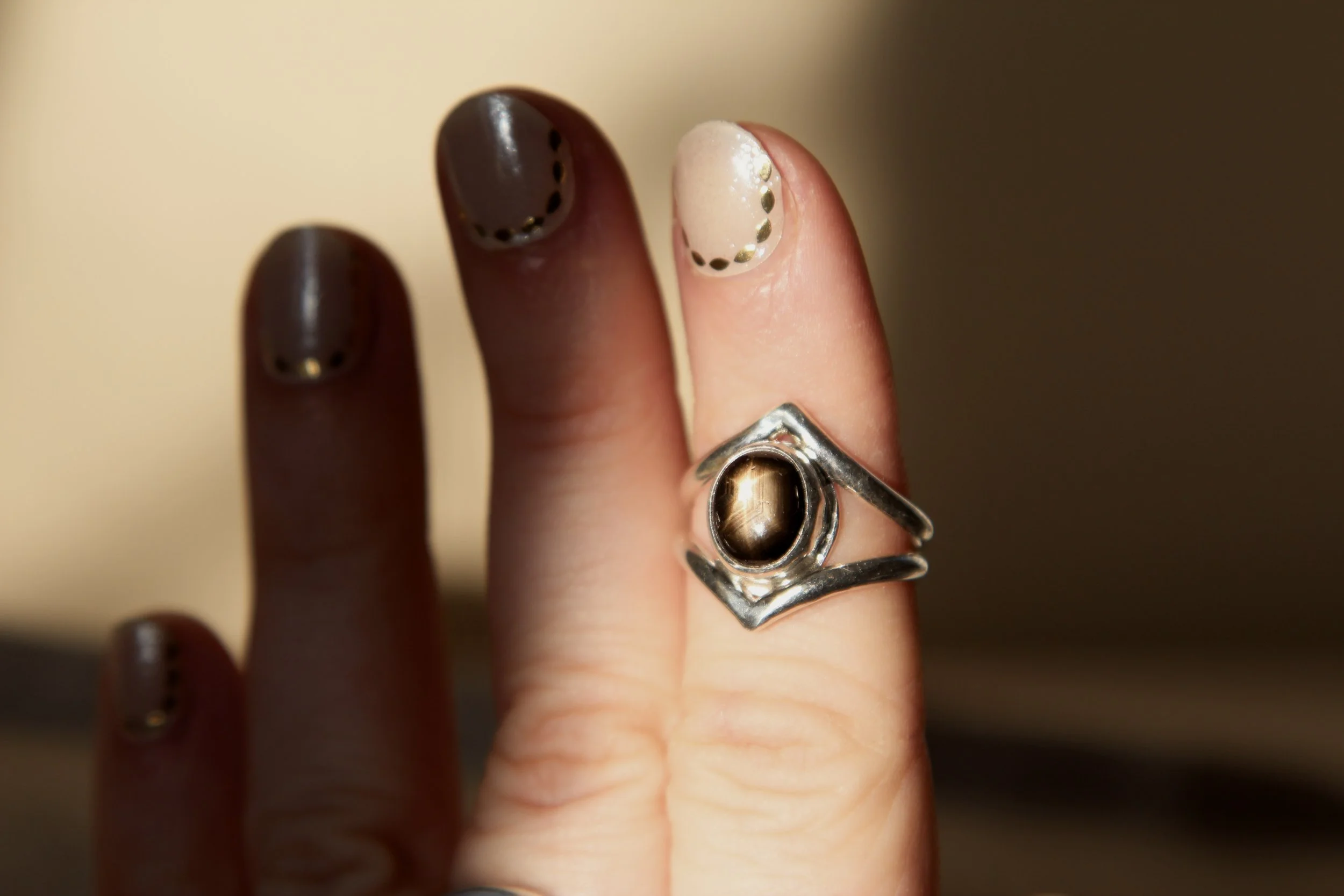 A hand with dark brown and white nail polish, decorated with dotted gold accents, wearing a silver ring with a black stone, against a neutral background.