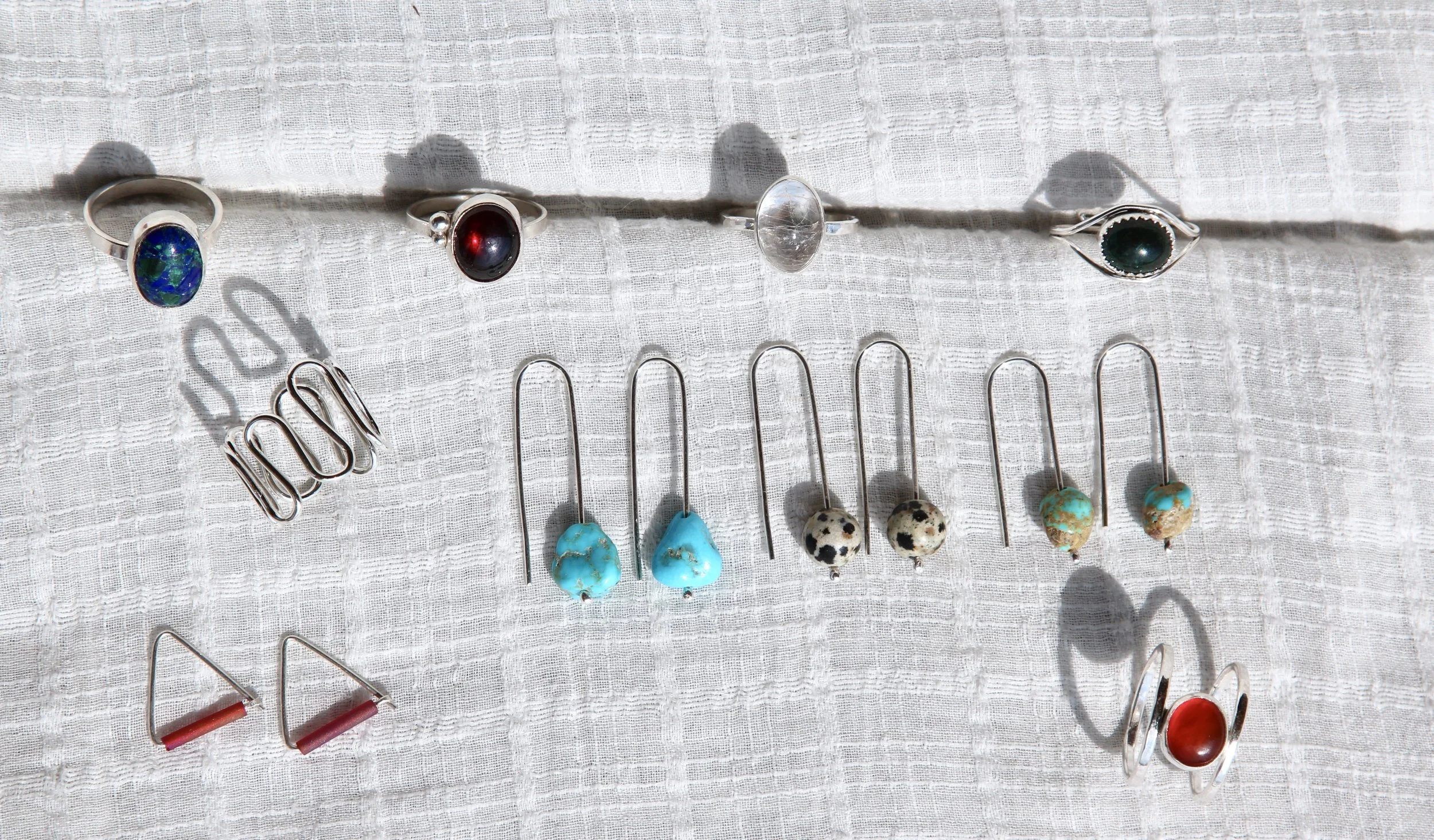 Assorted silver jewelry and earrings on a white textured cloth, including rings, a hair clip, and drop earrings with various stones and colors.