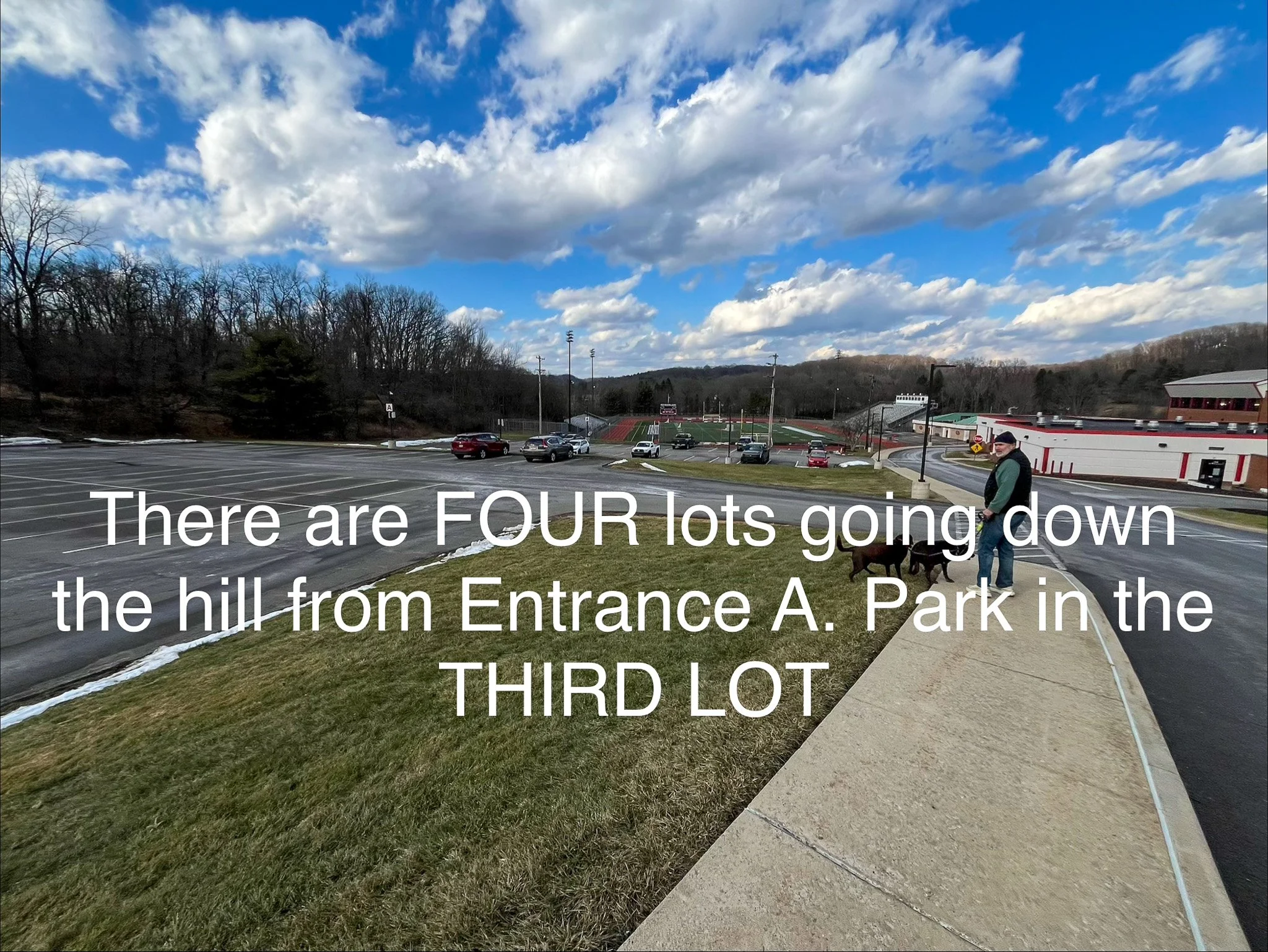 grass with a concrete sidewalk next to a parking lot with a man walking a dog and a blue sky with clouds. The words "There are FOUR lots going down the hill from Entrance A. Park in the THIRD LOT" over top