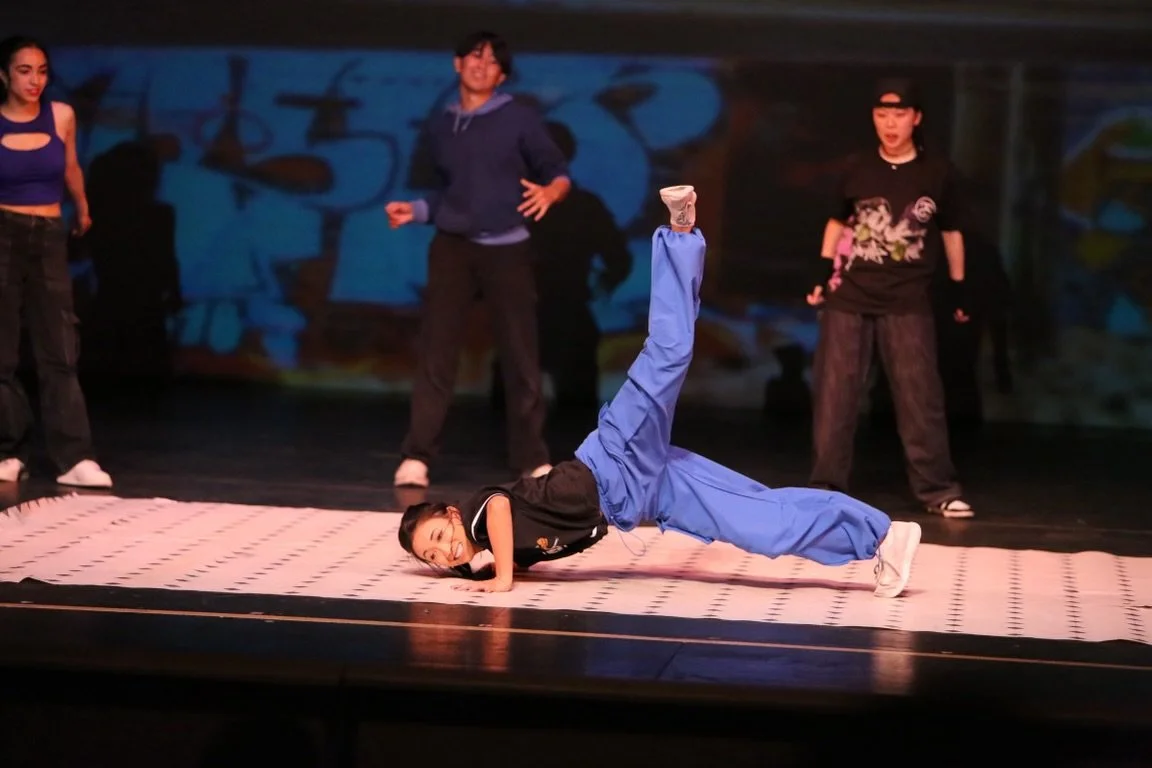 A woman with dark curly hair, wearing a black leather jacket, black top, and black pants, smiling and standing against a light purple background.
