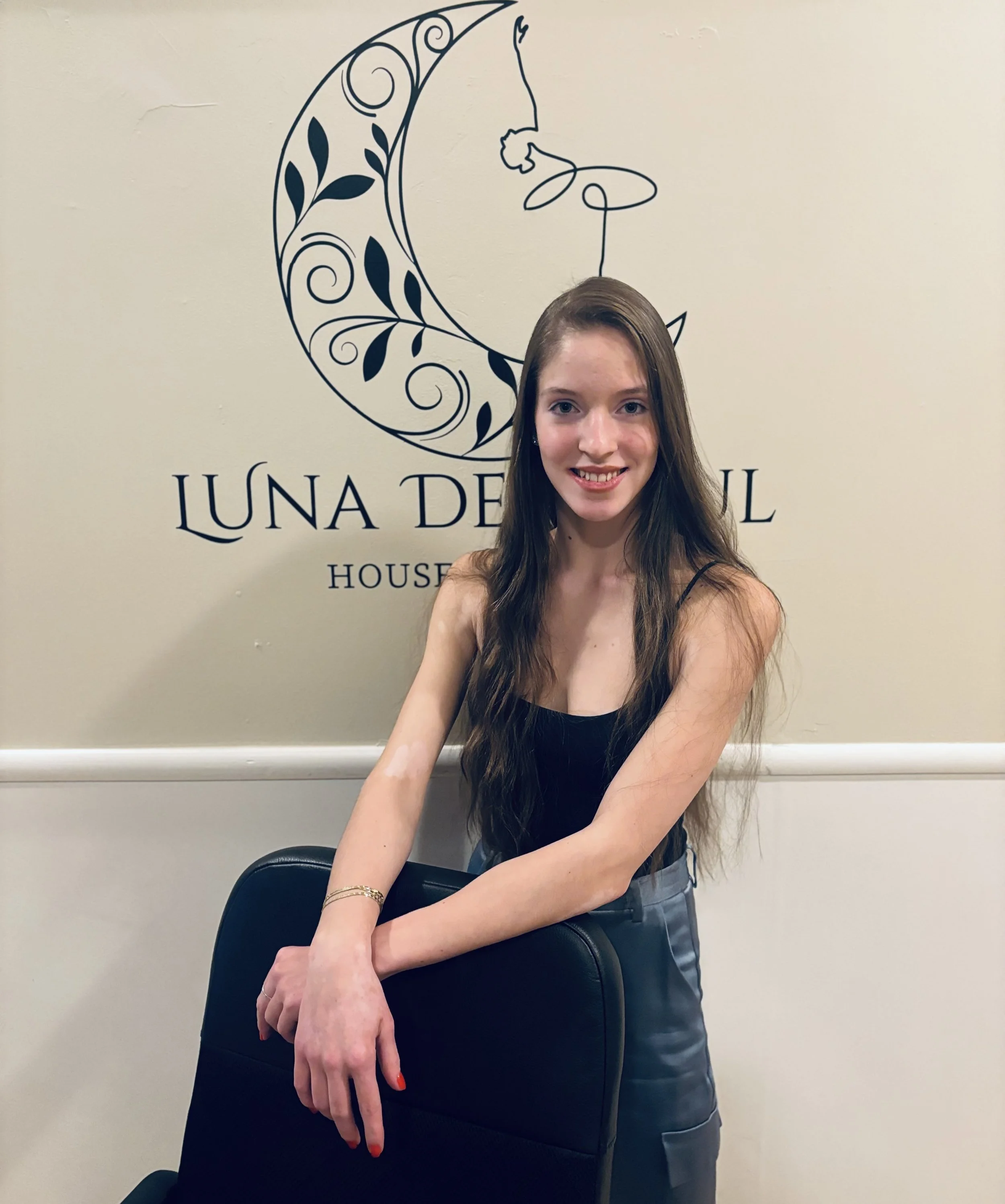 A man and woman standing close together on a wooden floor, smiling at the camera. They are dressed in black dance outfits, with the woman in a crop top and skirt, and the man in a long-sleeve shirt and trousers.