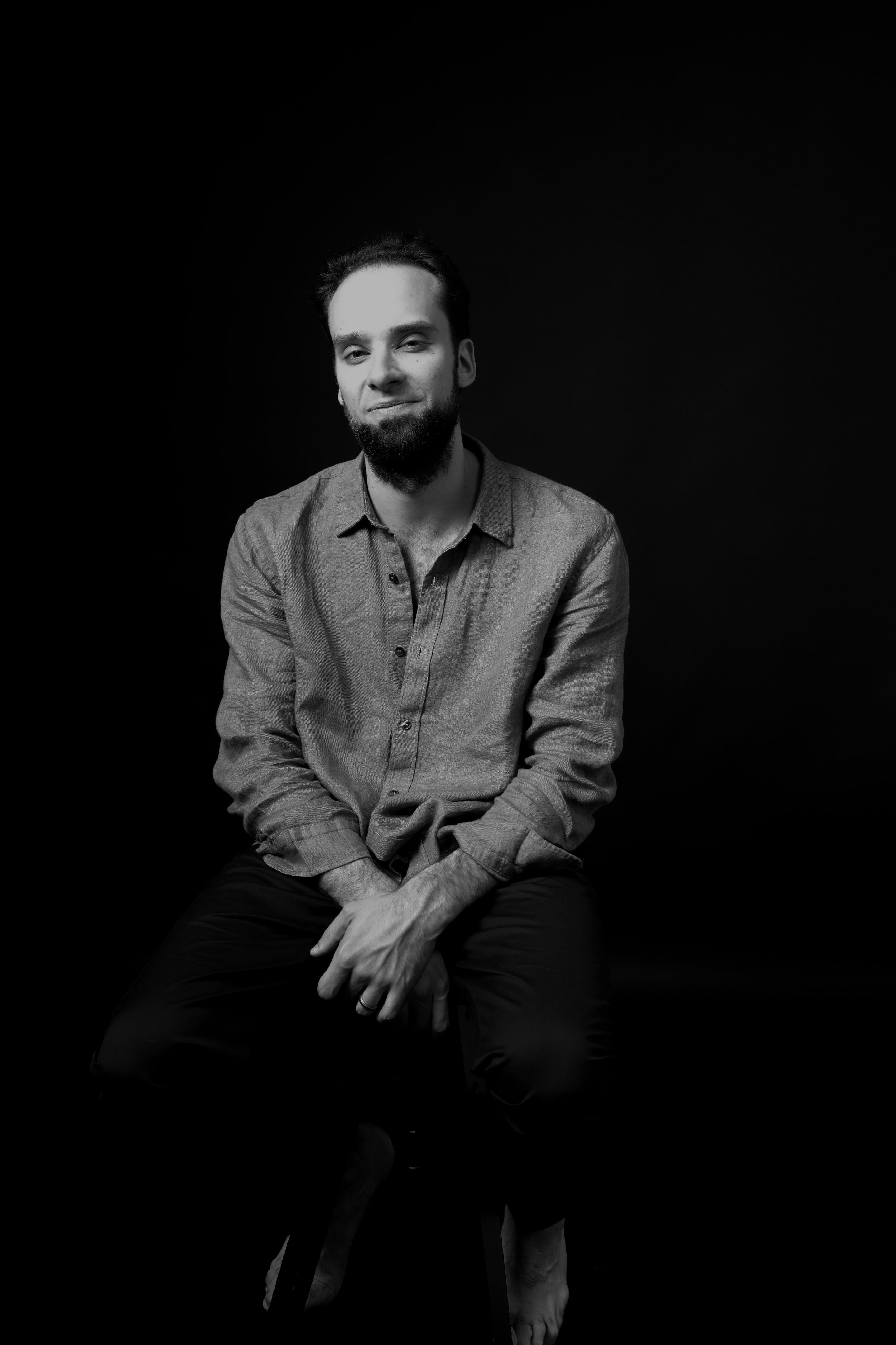 Black and white portrait of a man sitting on a stool against a dark background, wearing a button-up shirt and pants, with a relaxed posture and a slight smile.