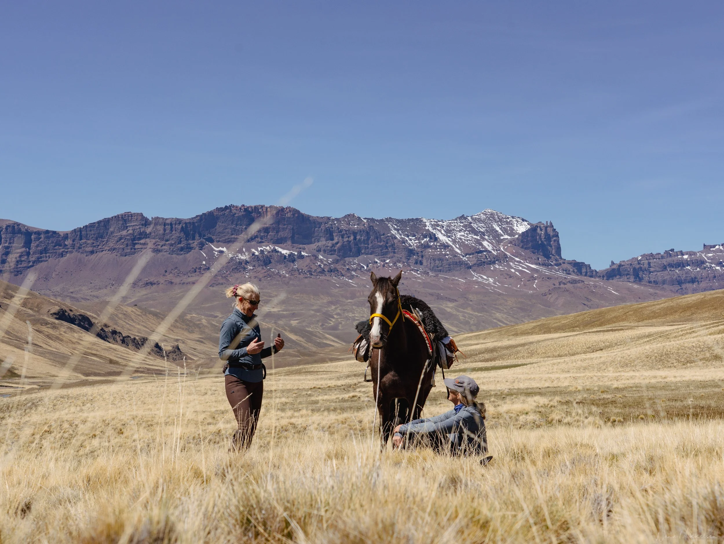 Two women taking a break with a Criollo horse in the highlands of Sierra Baguales, Chilean Patagonia, surrounded by open grasslands and rugged mountain landscapes.