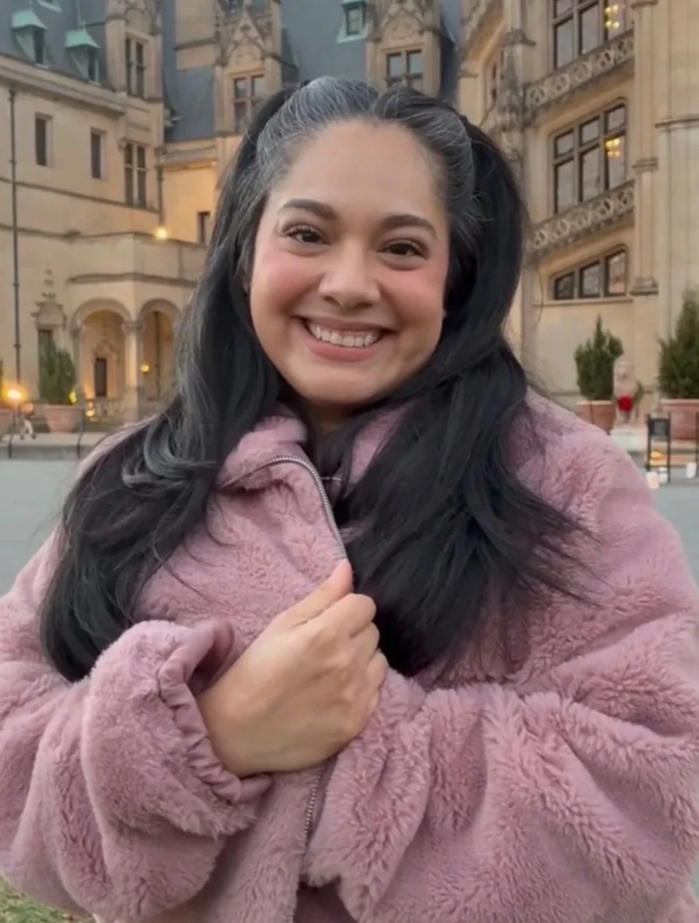 A young woman with long dark hair, smiling, wearing a pink fleece jacket, standing outdoors in front of a historic building with ornate architecture.