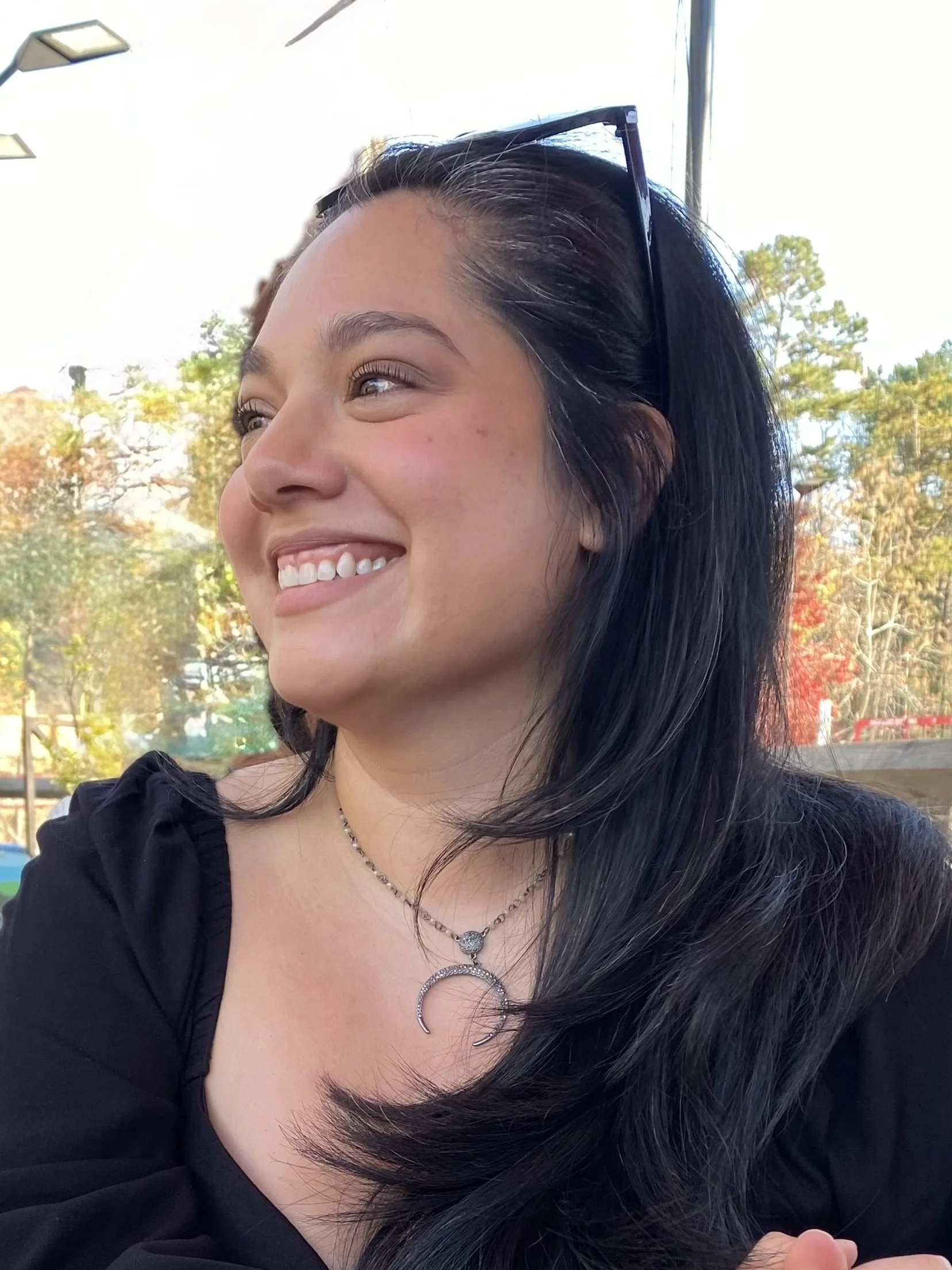 A young woman with black hair and sunglasses on her head, smiling outdoors with trees and a blue sky in the background.
