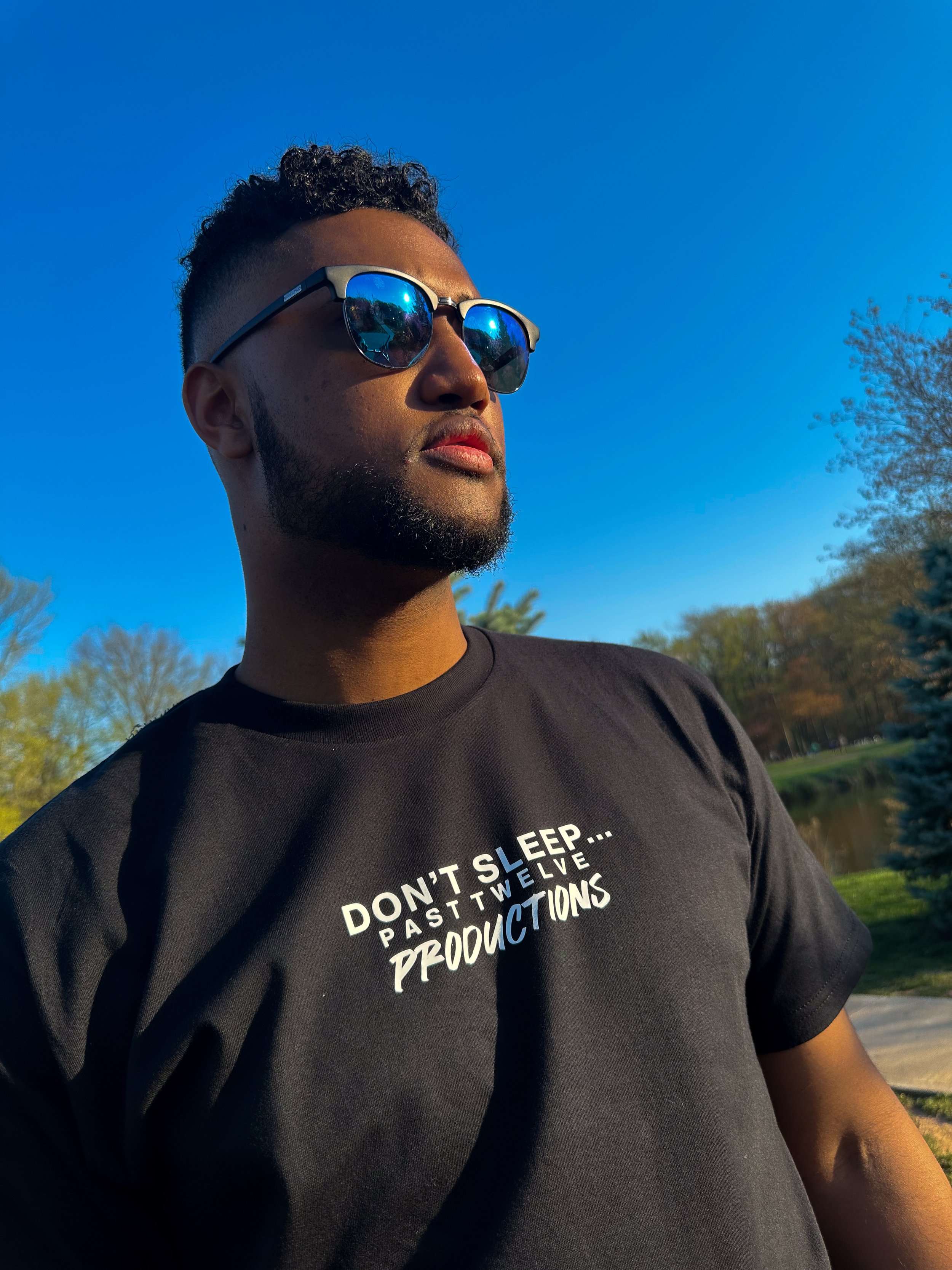 A man wearing reflective sunglasses and a black t-shirt with white and blue text in an outdoor park setting with clear blue sky and trees in the background.