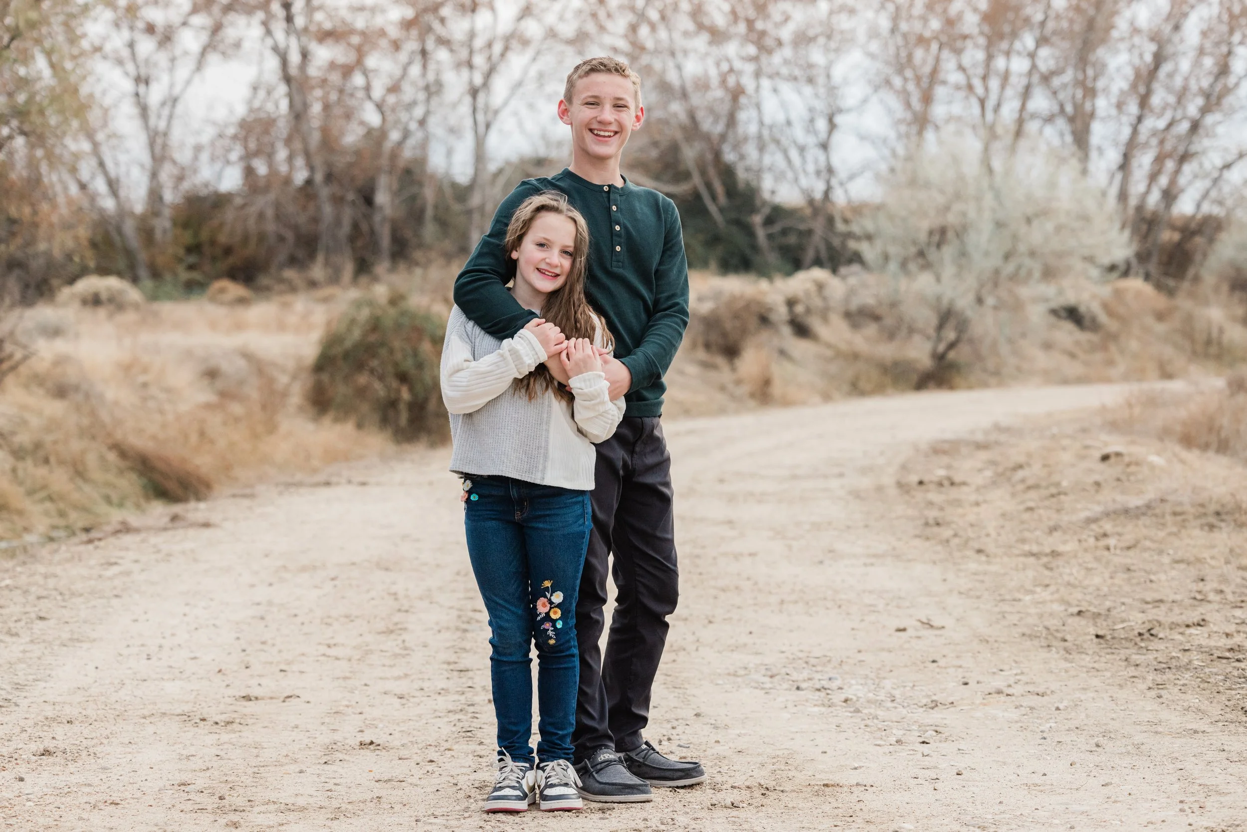 A smiling boy and girl stand together on a dirt path outdoors, holding hands, with trees and shrubs in the background.