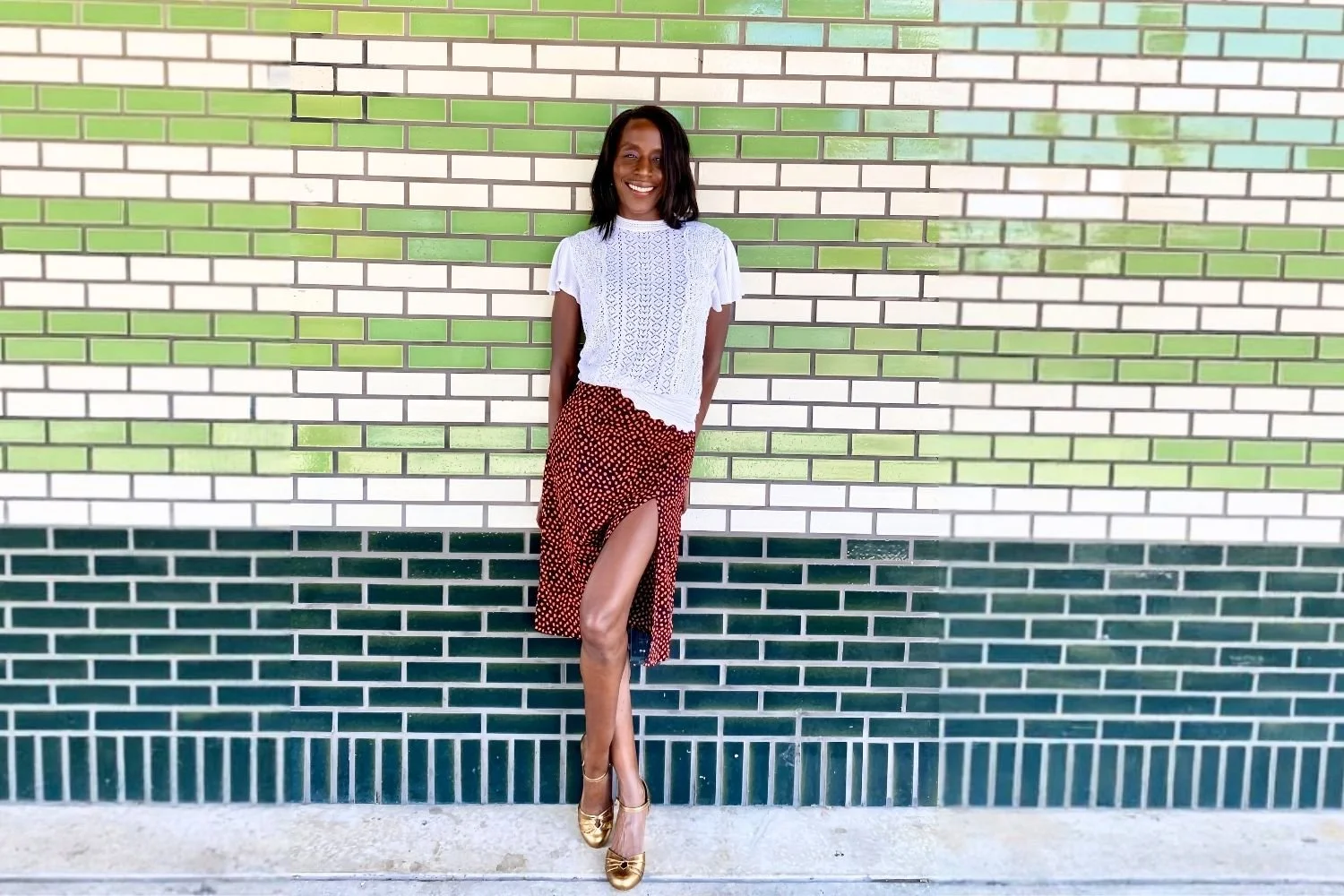 Margarette Allyn, author of Running from Yesterday, smiling and standing against a green and white tiled wall, wearing a white lace top, a rust-red polka dot midi skirt with a side slit, and gold heels