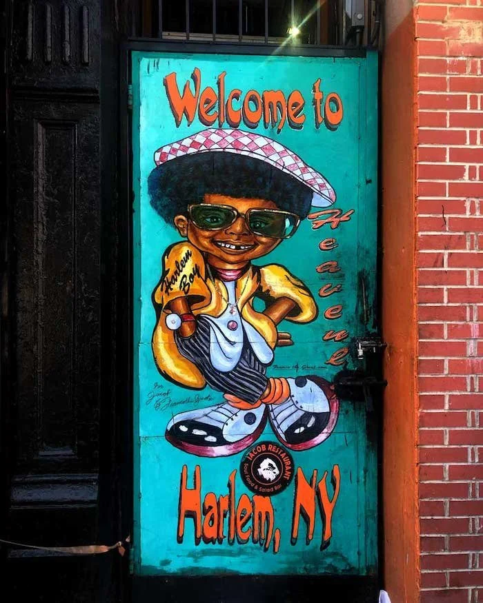 Colorful street mural on a teal door reading Welcome to Heaven Harlem NY featuring a Harlem Boy cartoon character in Harlem New York