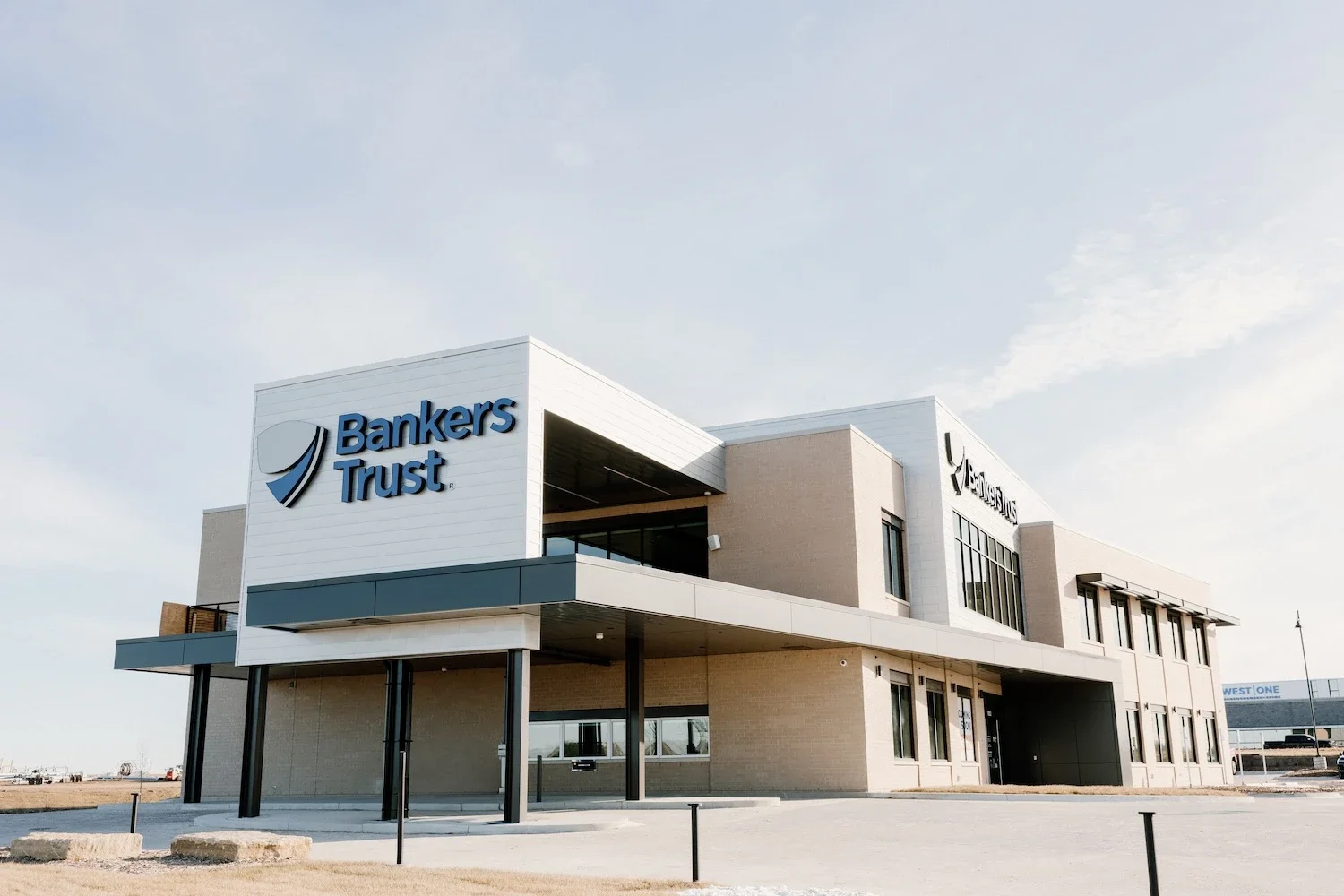 A modern two-story building with white and beige exterior walls, large windows, and signage that reads 'Bankers Trust' in blue letters, located in a commercial area under a clear sky.