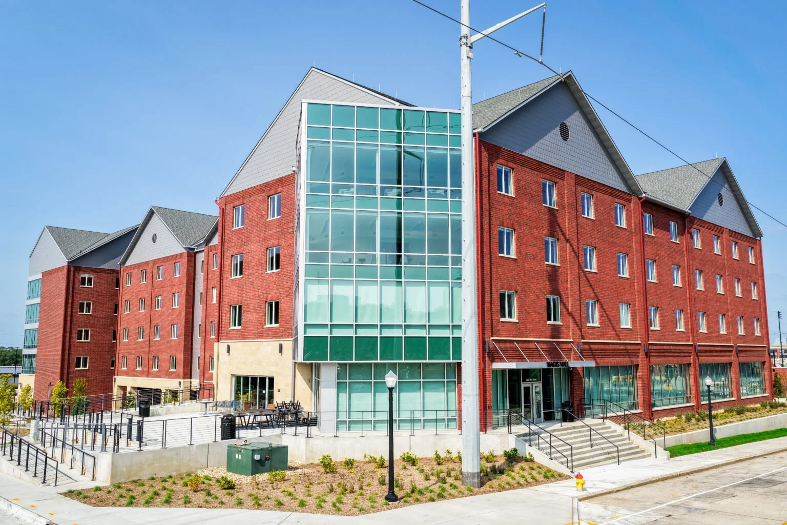 A modern multi-story apartment building with red brick exterior, gray gabled roofs, and large glass windows, located in an urban area with a sidewalk, street lamps, and landscaped surroundings.