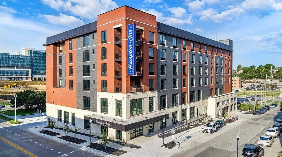 A modern mixed-use building with a Hampton Inn hotel on the upper floors and retail space on the ground level, located on a city street with parked cars and streetlights.