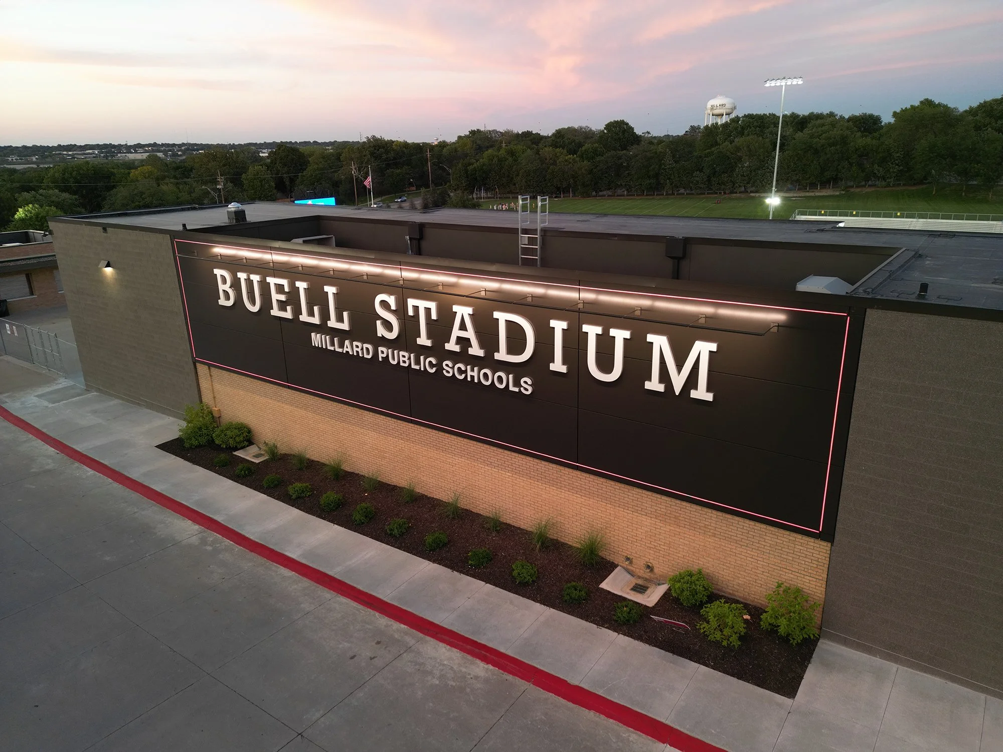 A school building with a sign that reads 'Buell Stadium Millard Public Schools' on the front facade, a running track, a flooded field, and a water tower in the background, during dusk with pink and blue sky.