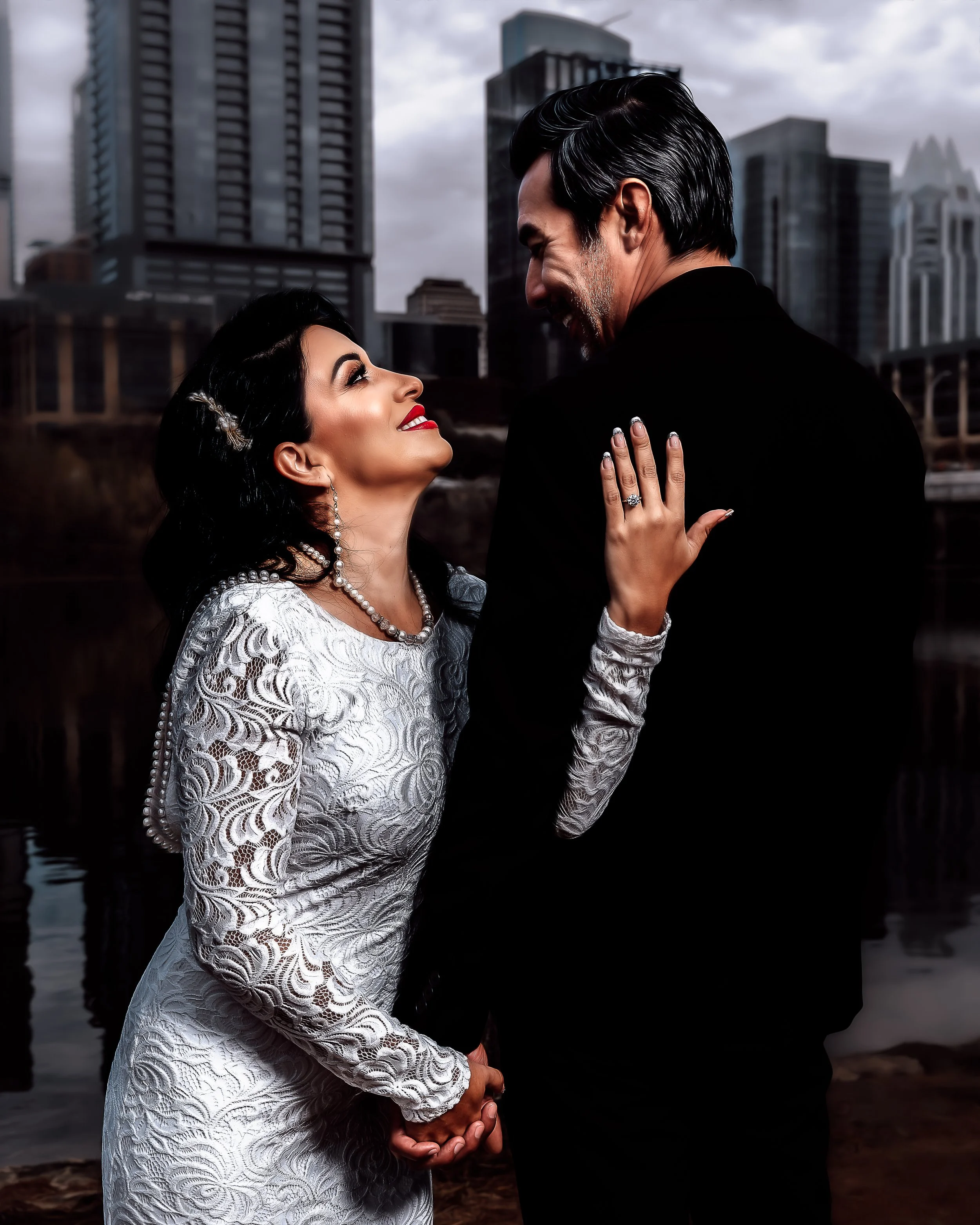 A couple dressed formally with a city skyline in the background. The woman is wearing a white lace dress and looking up at the man, who is in a dark suit.