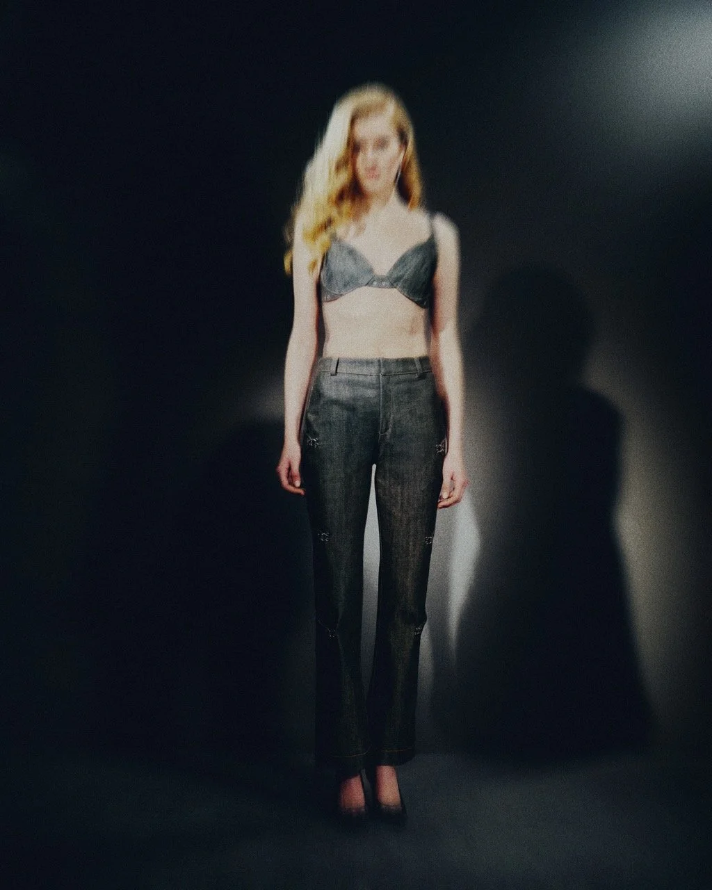 Blurry photo of a woman with long blonde hair wearing a black bra top and dark jeans, standing against a dark background with a shadow cast on the wall.
