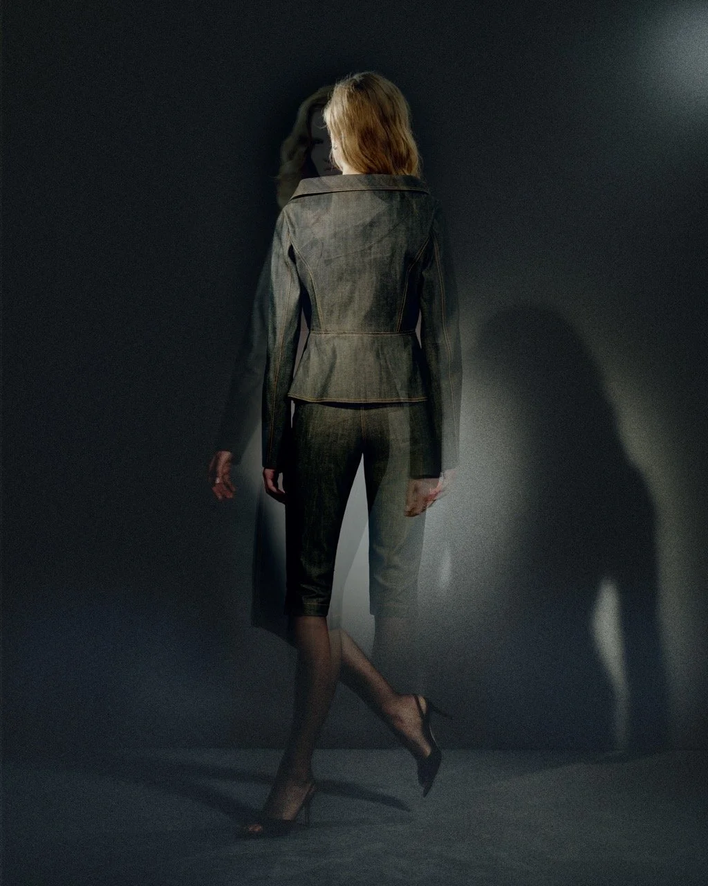 A woman stands facing away against a dark background, wearing a fitted suit with a blazer and cropped pants, and high heels.