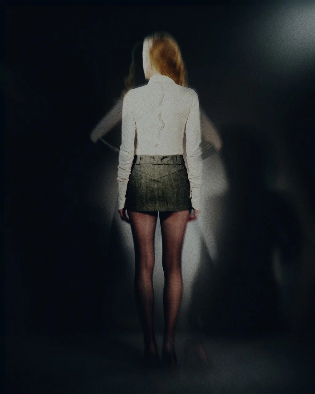 Woman seen from behind wearing a white blazer, dark plaid skirt, and sheer black tights, walking down a dimly lit hallway.