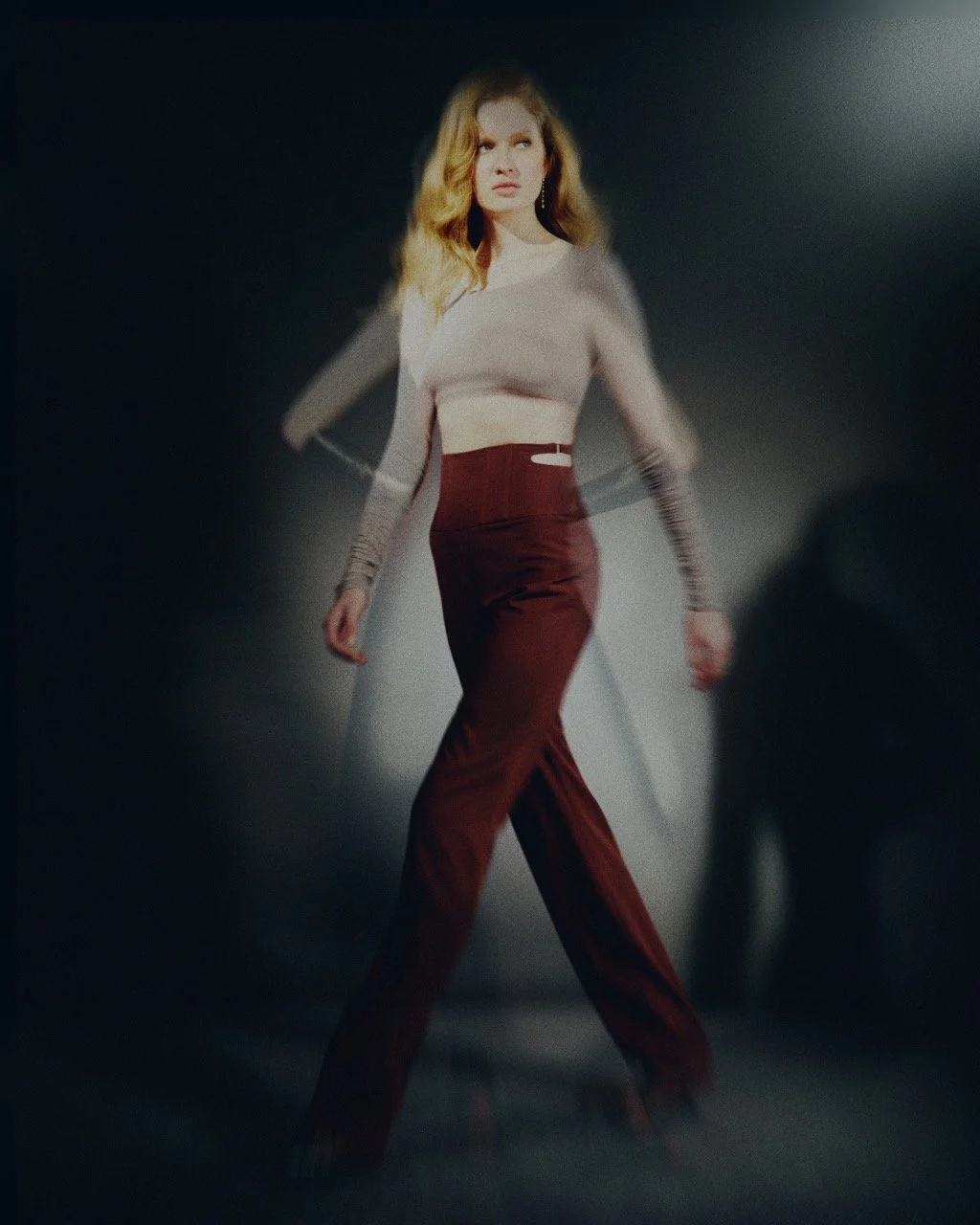 A woman with wavy blonde hair strikes a pose in a studio with a dark background, wearing a fitted light-colored crop top and high-waisted maroon pants.