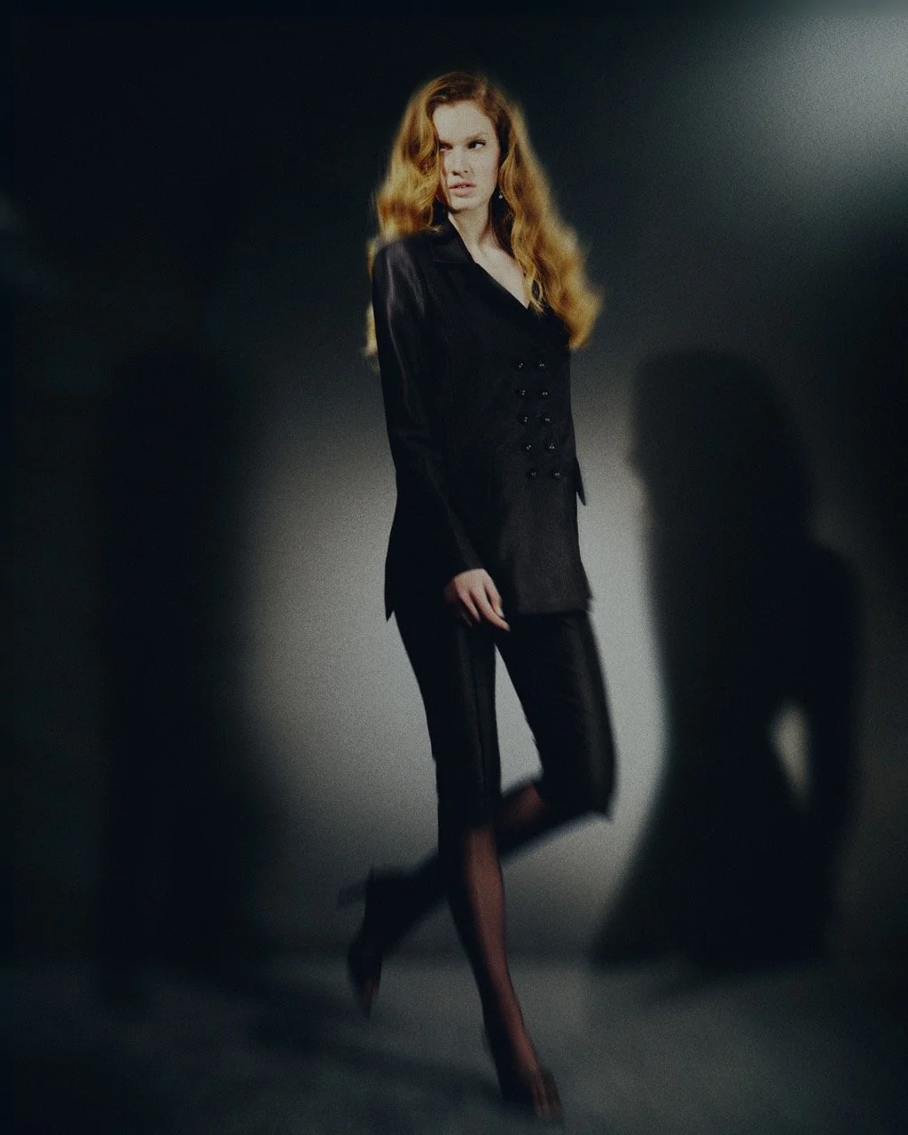 A woman with long red hair poses in a black outfit against a dark background with dramatic lighting.