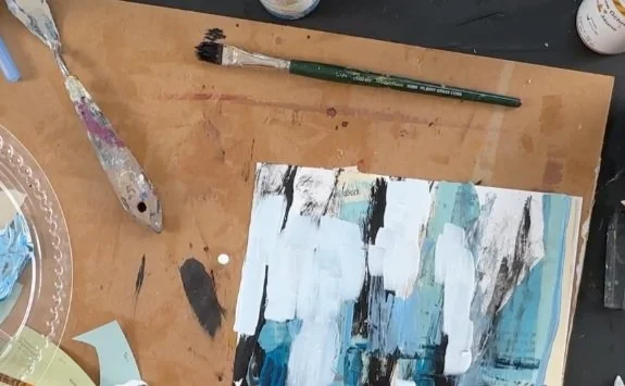 Abstract painting with blue, white, black, and brown brushstrokes on paper, surrounded by art supplies including paintbrushes, paint, and a palette knife on a wooden table.