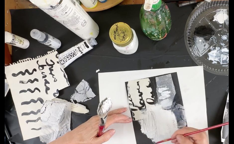 An artist is painting a black and white abstract picture with text on a table surrounded by paint bottles, brushes, a glass jar, and torn paper.