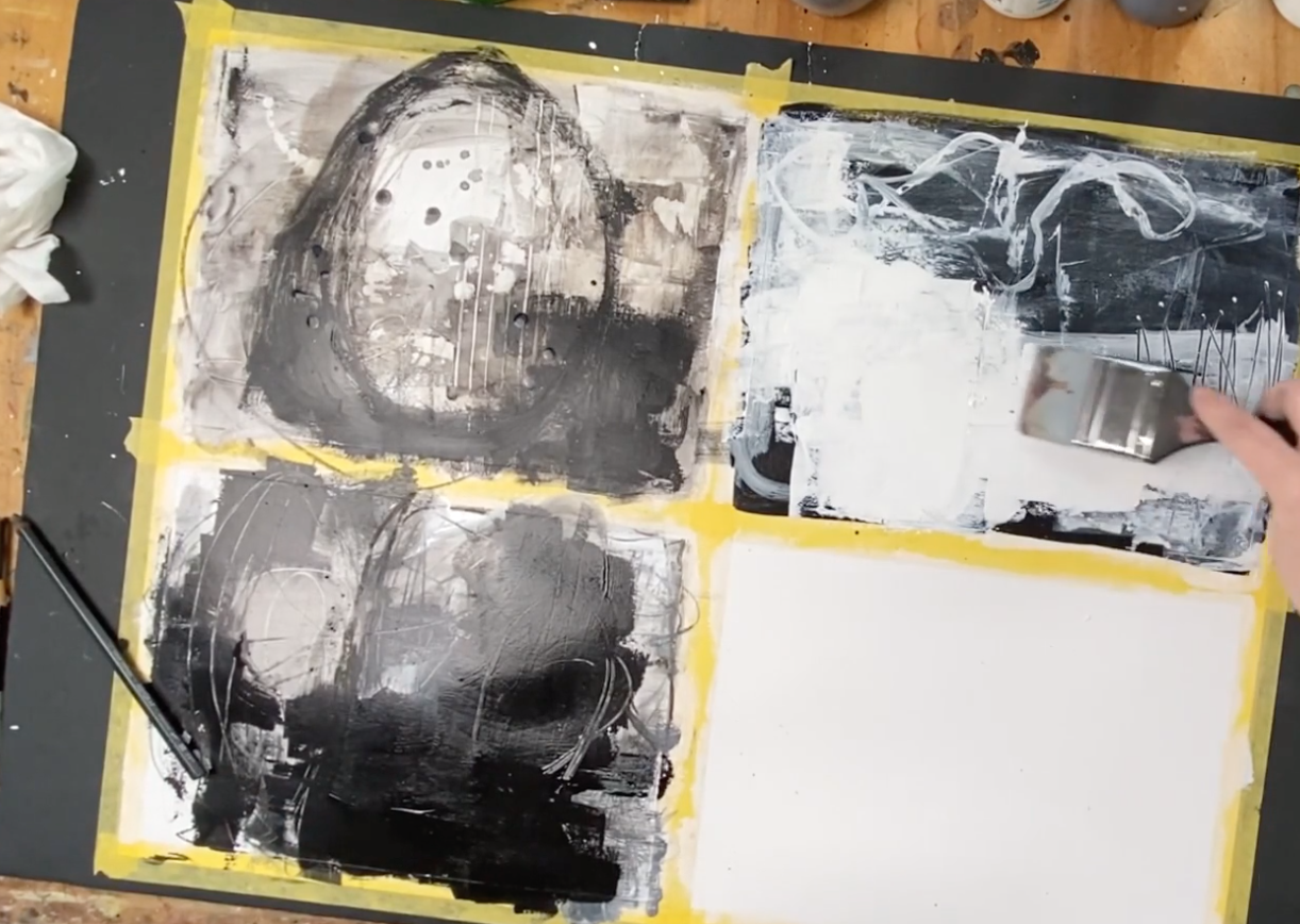 A person smearing white paint onto a mixed media canvas with black, white, and gray abstract designs, taped on a wooden table.