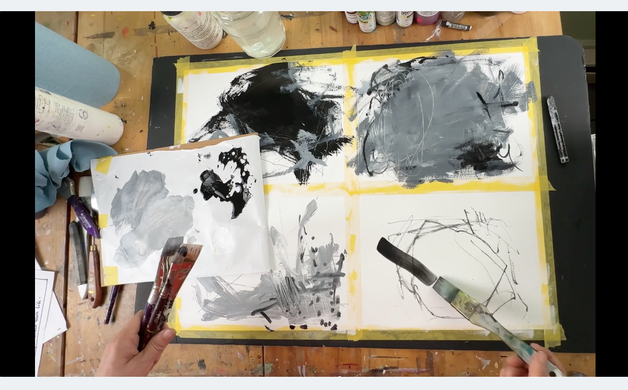 An artist's workspace with four abstract black and gray paintings on watercolor paper taped to a table, various paintbrushes, paint bottles, a palette, and art supplies visible around and below the papers.