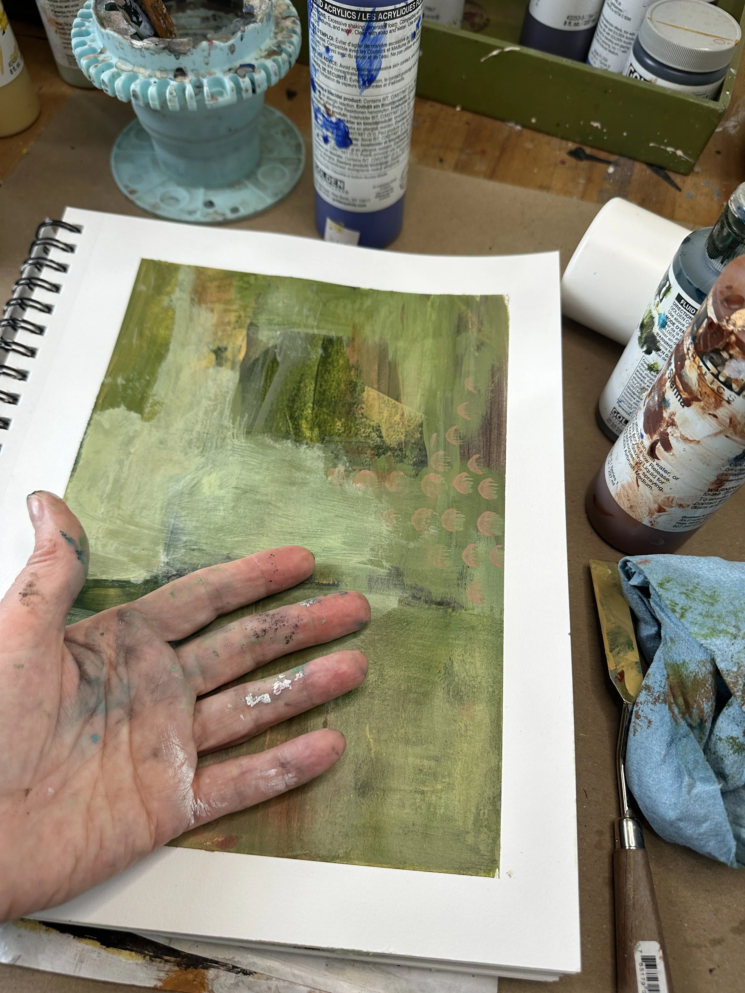 An artist's hand covered in paint rests on a landscape painting in progress, surrounded by various bottles of acrylic paint, a palette, and painting supplies on a wooden table.