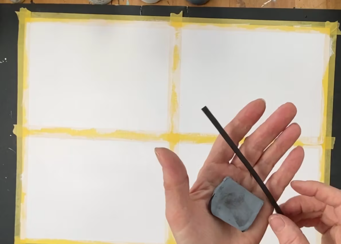 A hand holding a black strip of material and a square piece of grayish chalk or eraser, with taped white paper divided into four sections in the background.