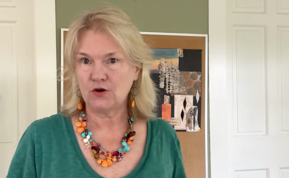 A middle-aged woman with blonde hair wearing a green shirt and colorful necklace and earrings, standing in front of a bulletin board with abstract art in a room with white walls and trim.