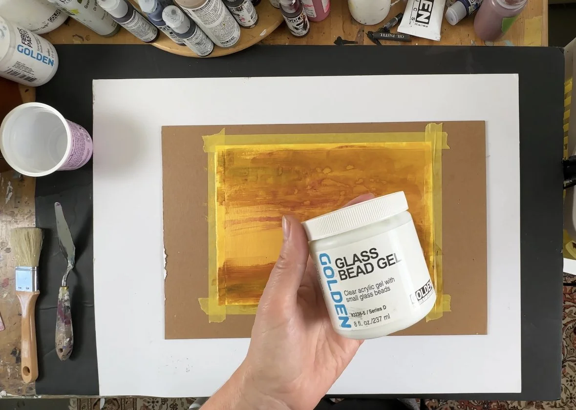 A person holding a container of golden glass bead gel over a canvas with painted horizontal streaks of orange, yellow, and red, taped to a work surface surrounded by art supplies.