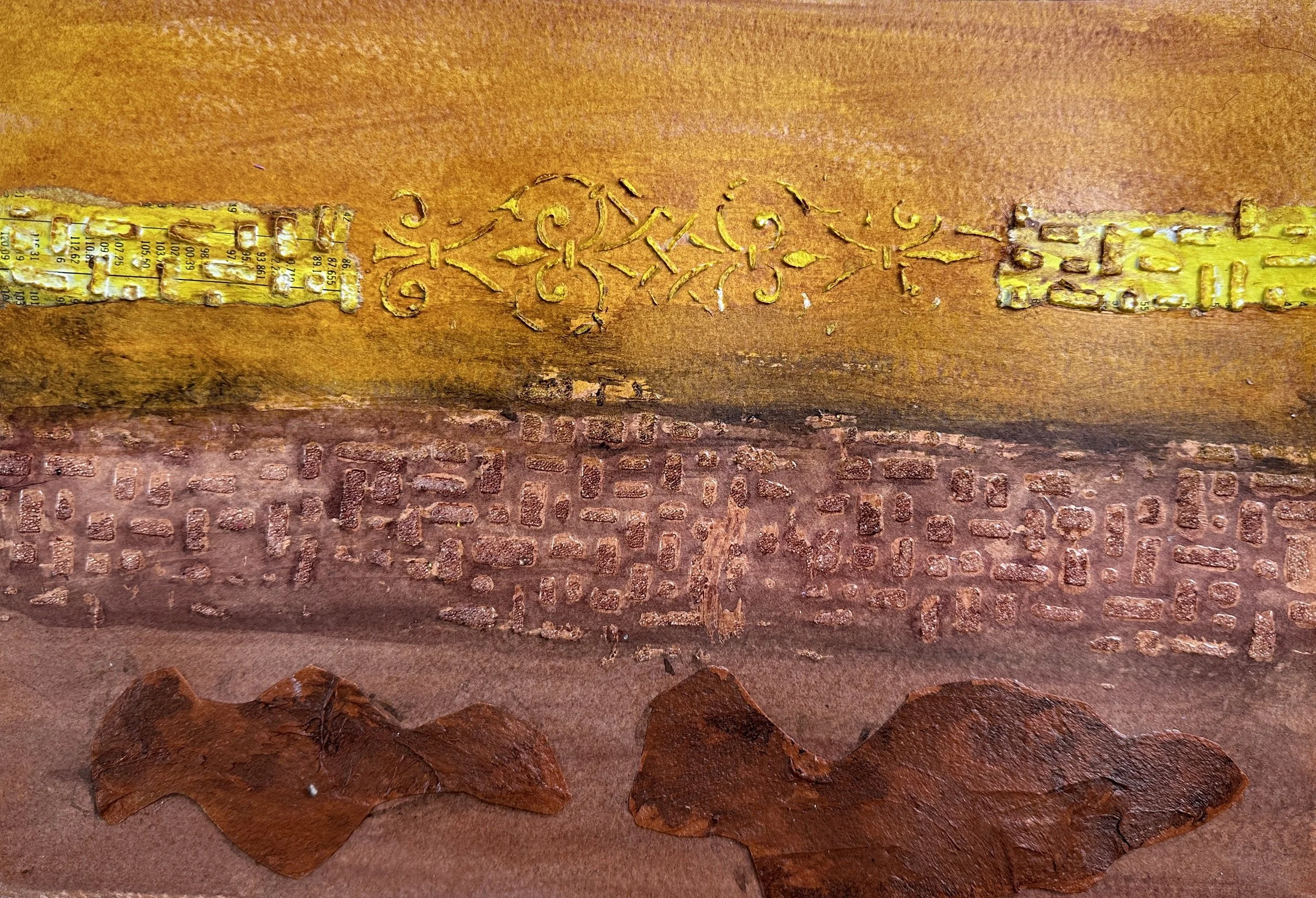 Close-up of a textured surface with a mix of materials, including a strip with measuring marks, decorative gold-colored patterns, a brick-like pattern, and two brown rocks.