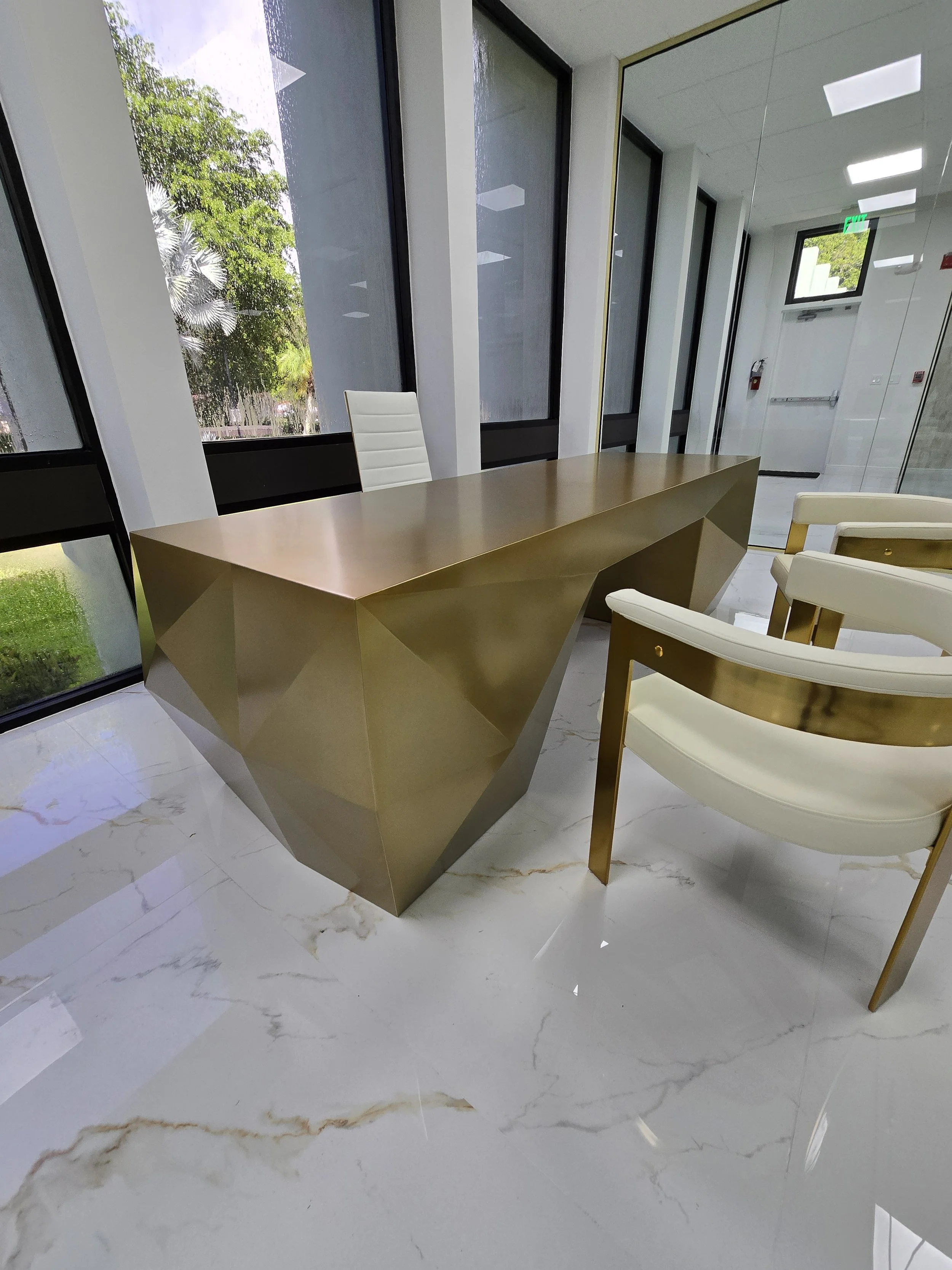 Modern office with a gold geometric desk, white leather executive chair, and cream upholstered guest chairs with gold accents, next to large windows and marble flooring.