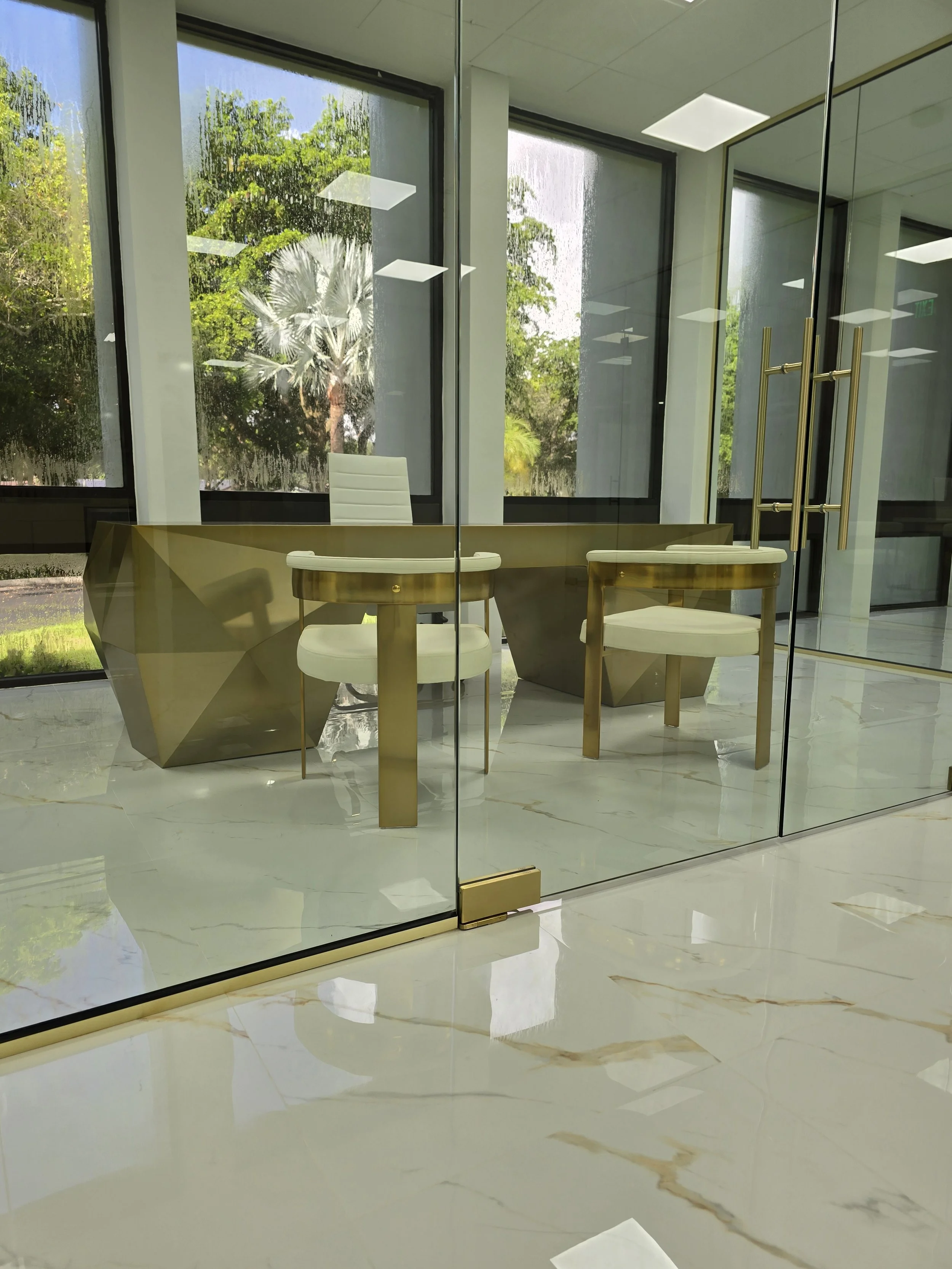 An office with gold and white furniture, including a desk and chairs, seen through a glass wall. Outside, green trees and a palm tree are visible.