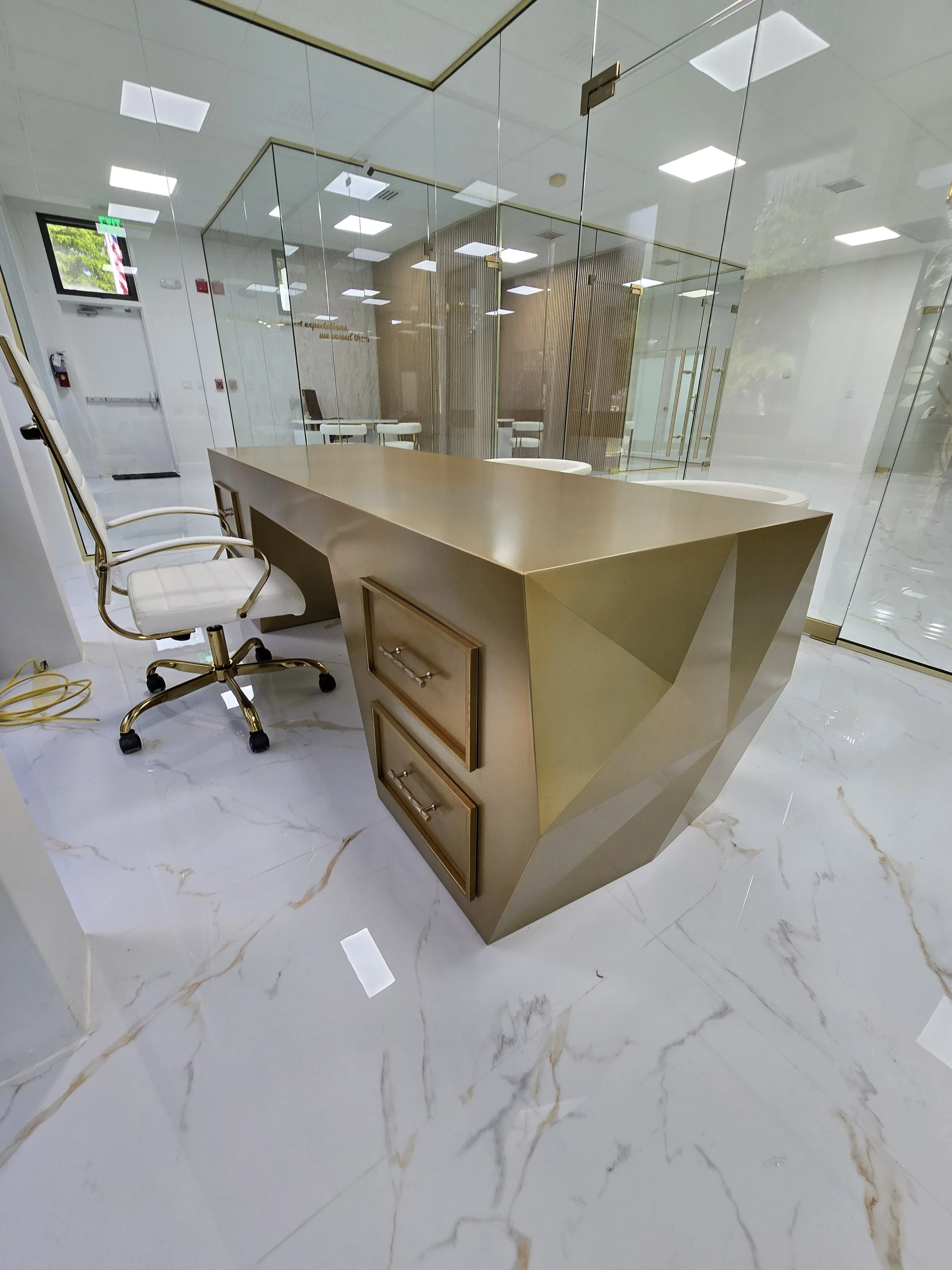 Modern office reception area with a gold geometric desk, white chairs with gold accents, and a glass wall with reflections.