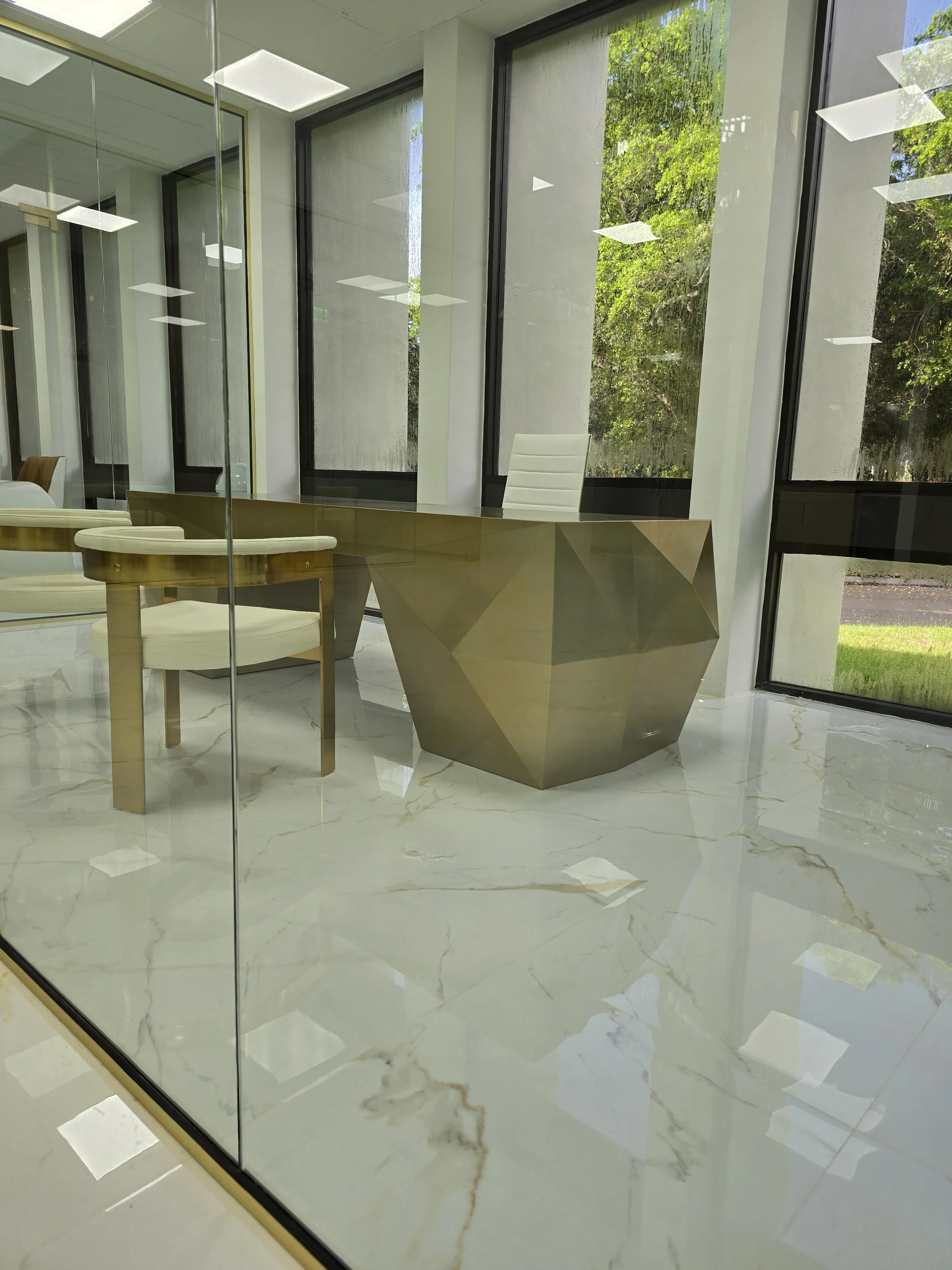 Modern office reception area with a geometric gold desk, a white office chair, and large glass windows showing greenery outside.