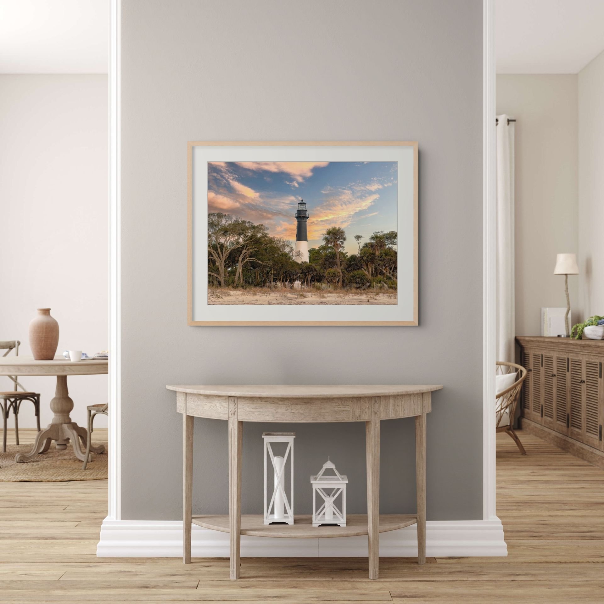 Interior of a modern home with a light gray wall and a framed picture of a lighthouse, trees, and a colorful sky. In front of the wall, there's a decorating table with lanterns on the floor, and decorative items on top. To the left, a wooden table with a ceramic vase and cups is visible. To the right, part of a basket and a table lamp can be seen.