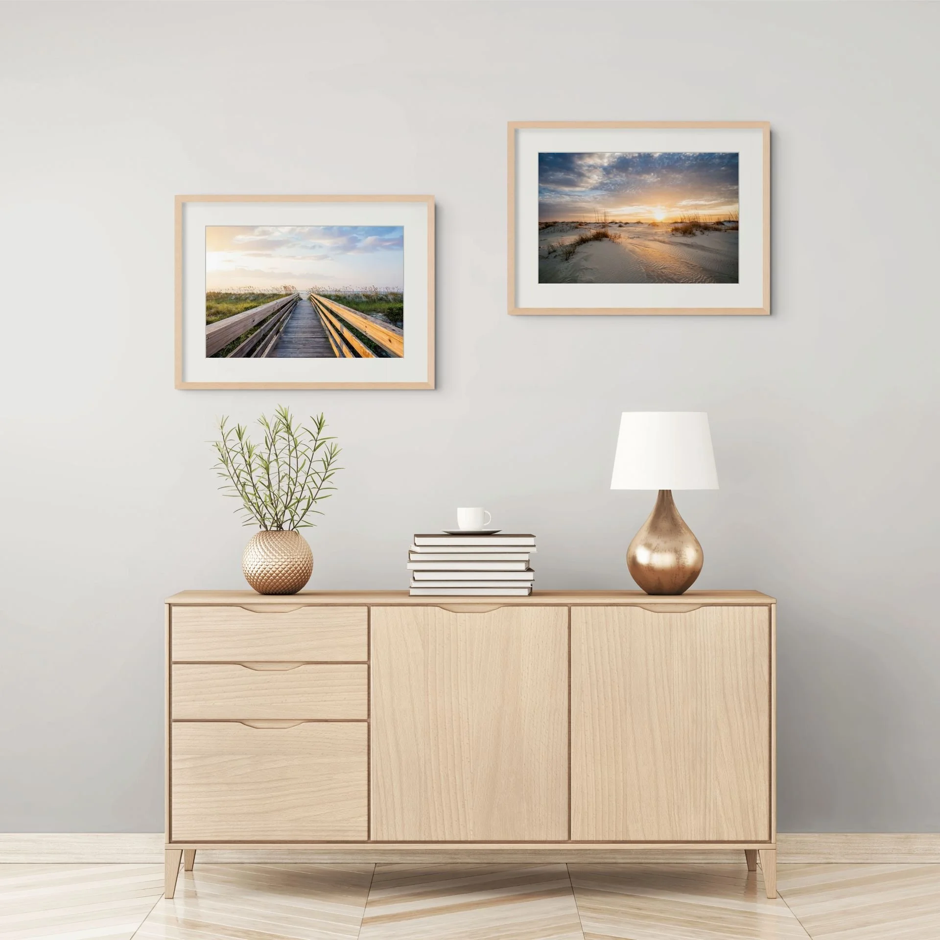 A wooden sideboard with decorative items, including a potted plant, a stack of books with a cup on top, and a metallic vase. Two framed landscape photographs of beaches and sunsets are hung on the white wall above. A table lamp is partially visible on the right.