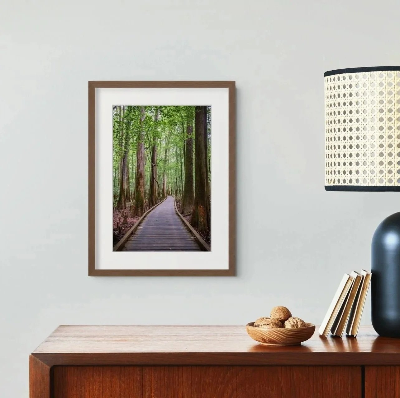 A framed photograph of a forest trail hangs on a white wall above a wooden sideboard. On the sideboard are a bowl of walnuts, a few books, and a black table lamp with a white rattan-patterned shade.