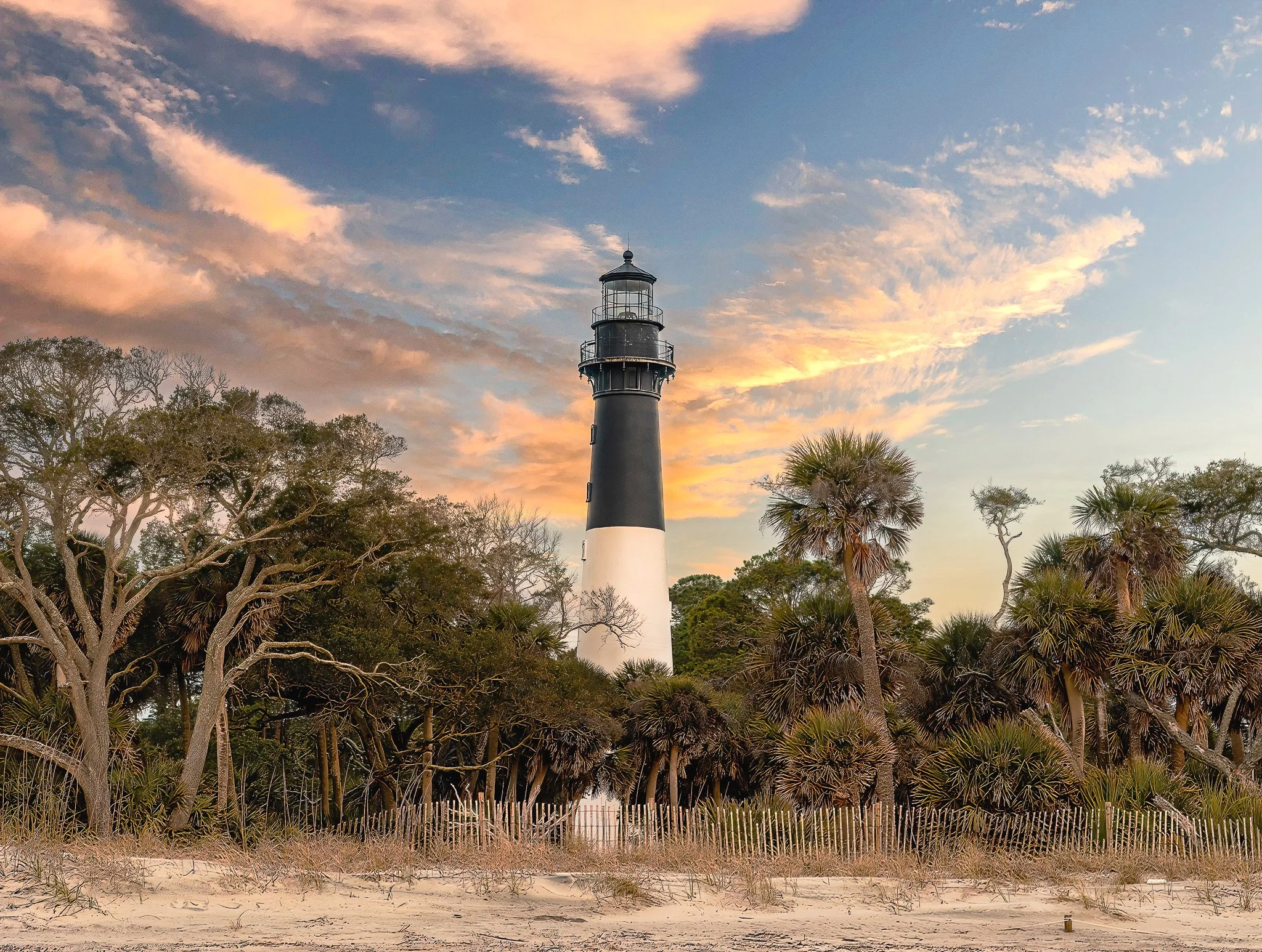 A lighthouse standing among trees and sand dunes during sunset with a sky filled with pink and orange clouds.