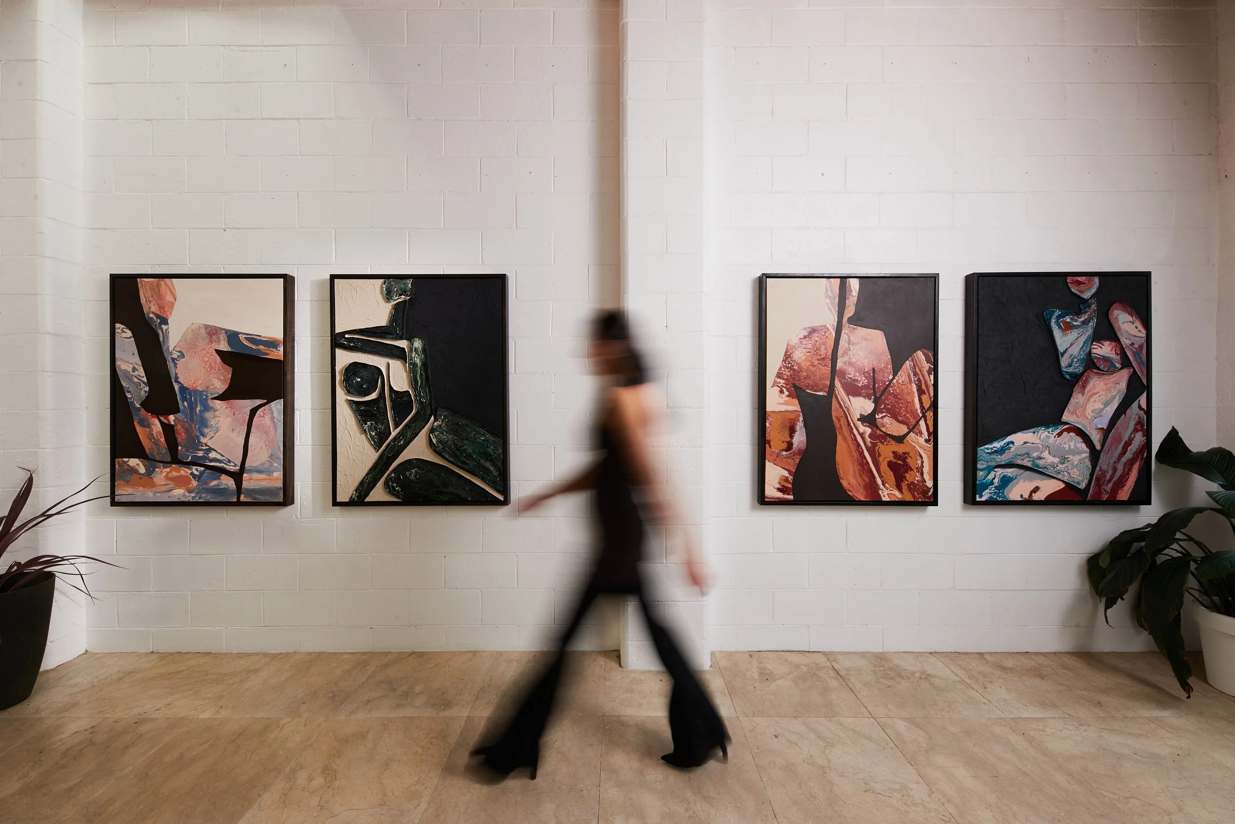 A person walking past four abstract paintings hung on a white brick wall in an art gallery.