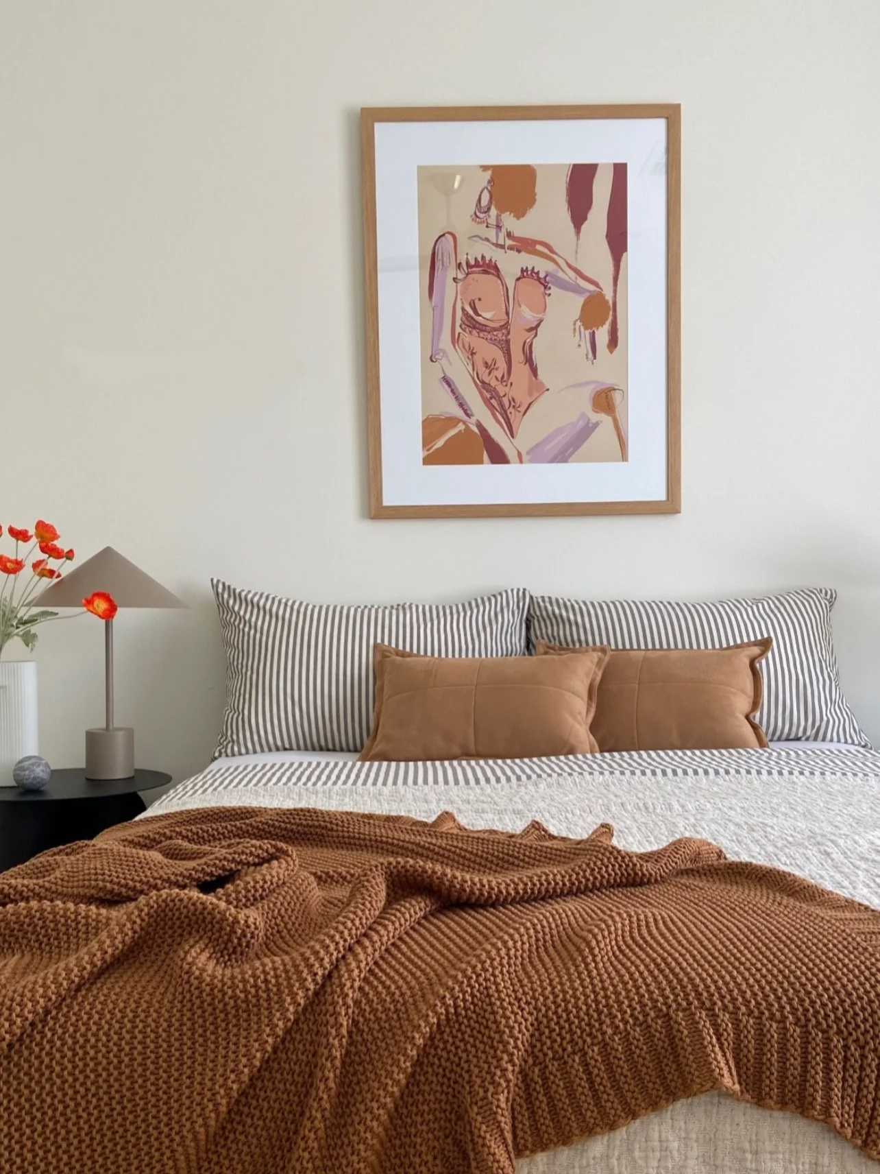 A cozy bedroom with a bed that has striped pillows, a brown throw blanket, and two brown pillows. There's a nightstand with a lamp, a vase with red flowers, and a small decorative object. A large abstract art piece hangs above the bed, and the walls 