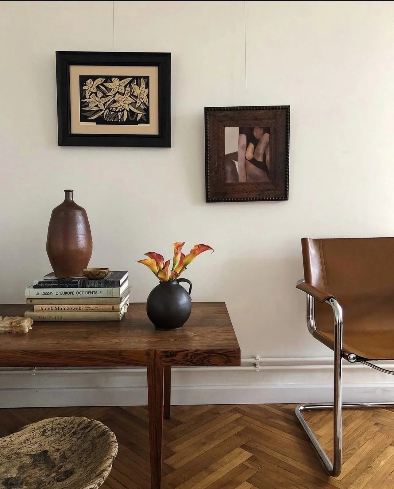 Interior of a room with a wooden table holding a brown vase with calla lilies, stacked books, and a small dish. Wall art includes a painting of yellow flowers in a black frame and an abstract photo in a dark wooden frame. A leather and metal chair is