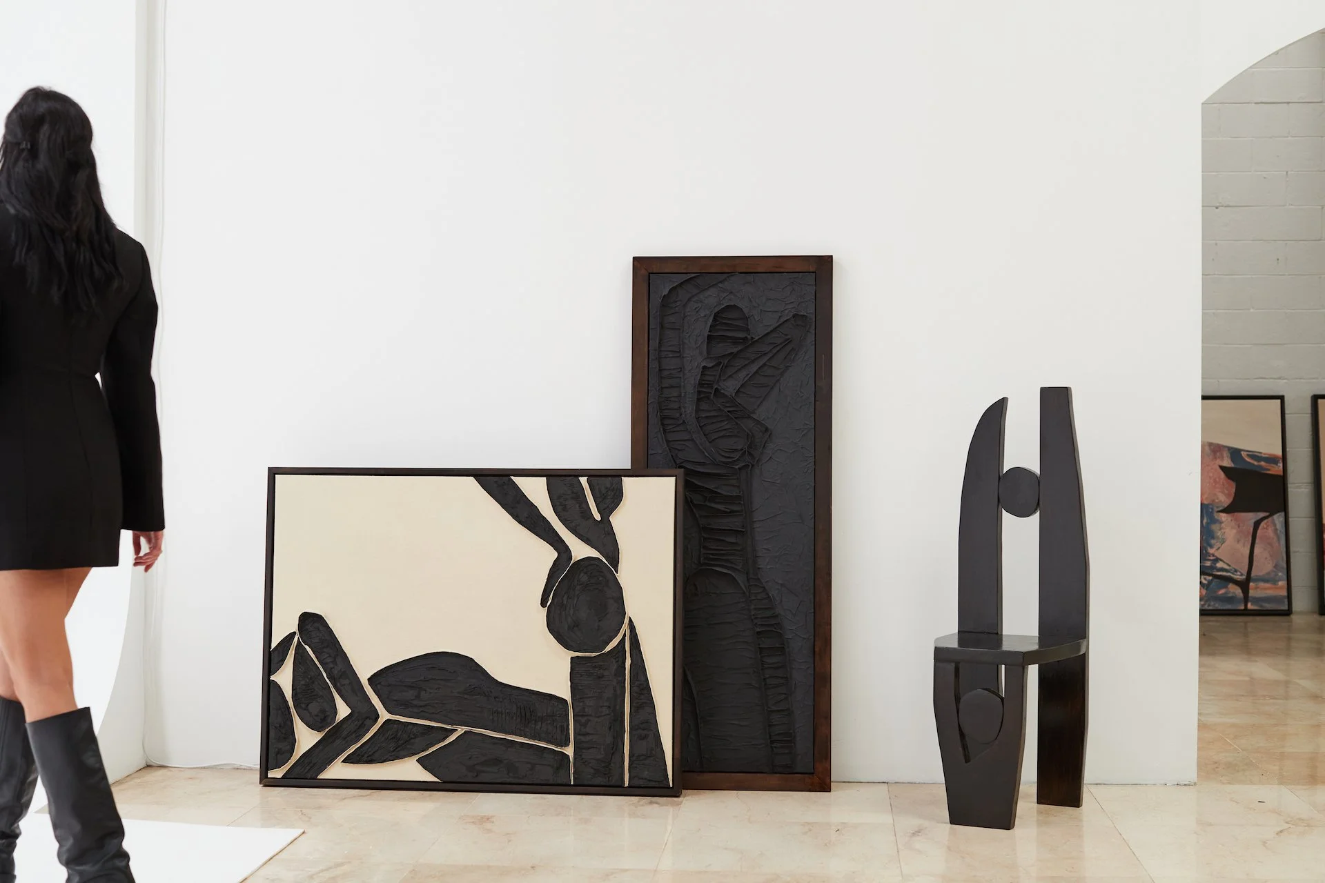 Art gallery with abstract black and beige paintings, a black sculpture, and a woman in a black coat and boots observing the artwork.