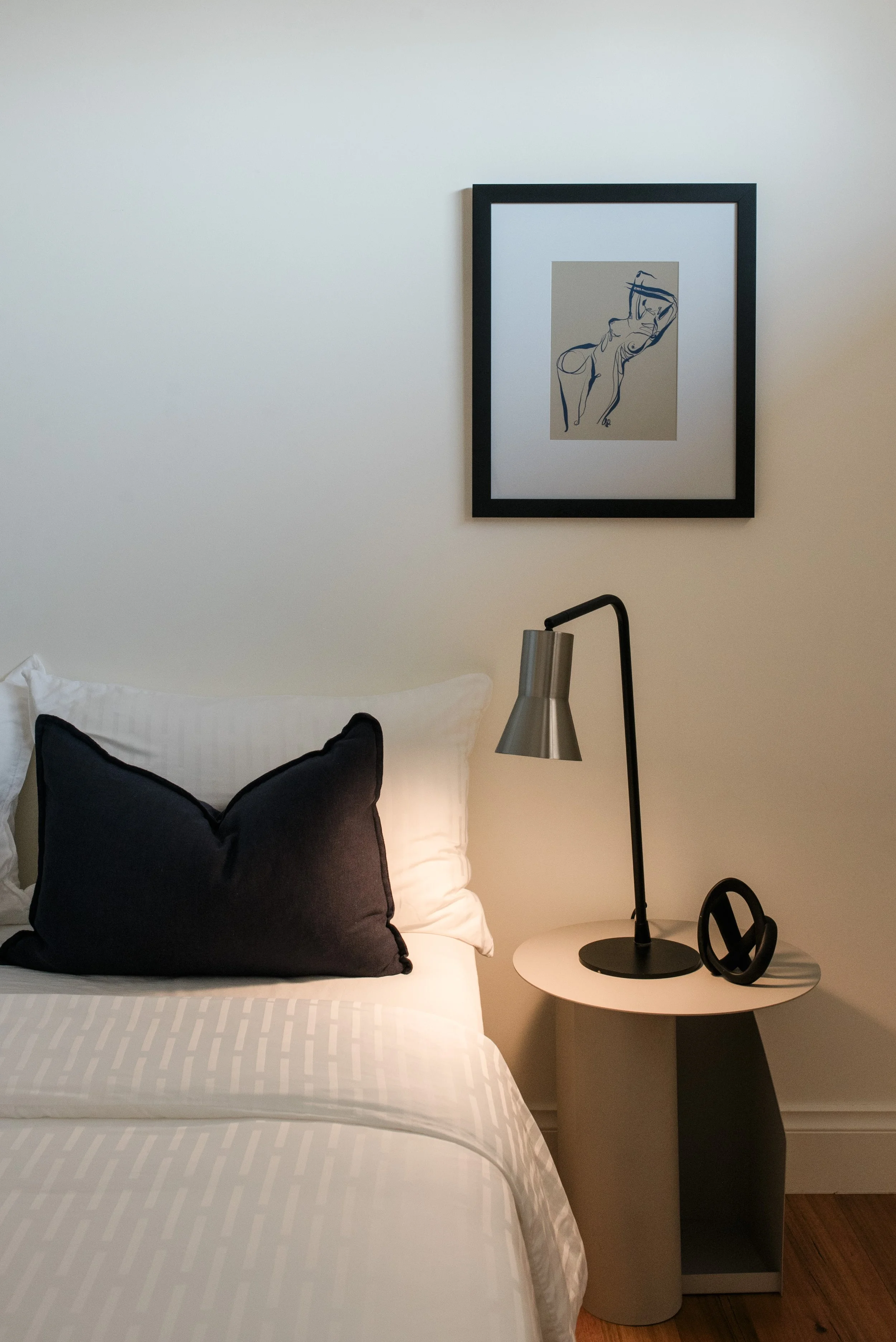 A neatly made bed with white and dark blue pillows, a bedside table with a modern lamp, and a framed line art picture of a woman hanging on a plain wall.