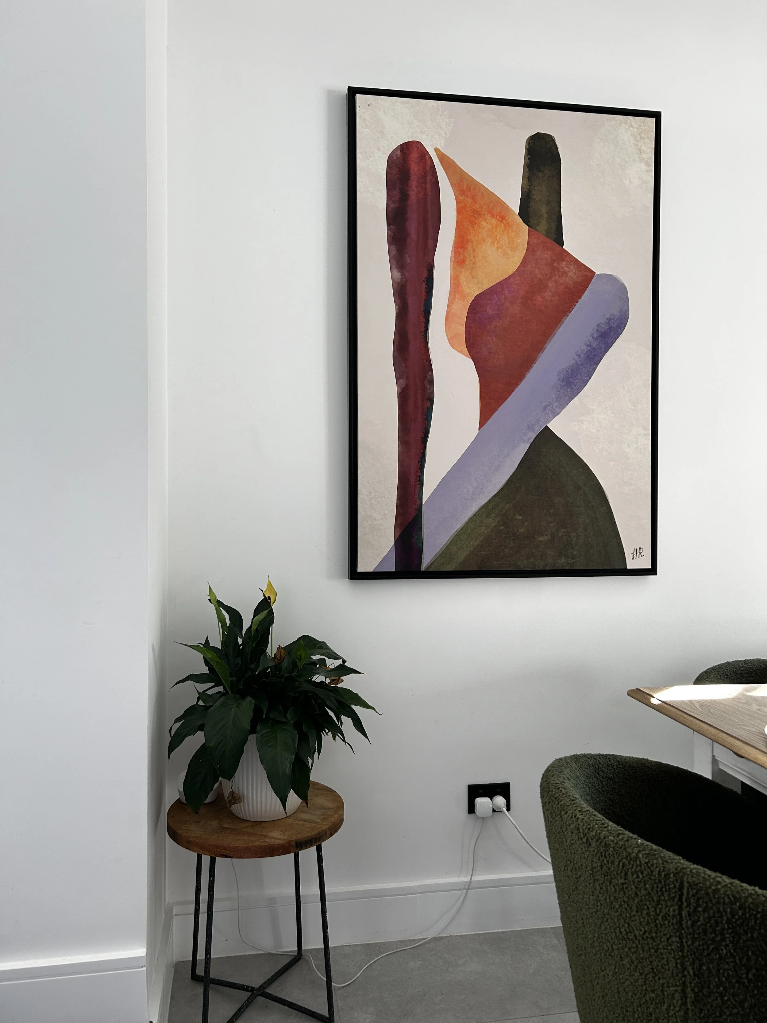 Interior of a modern room with a framed abstract art piece on the wall, a potted plant on a small round wooden table, and a partially visible green upholstered chair near a wooden table with sunlight streaming in.