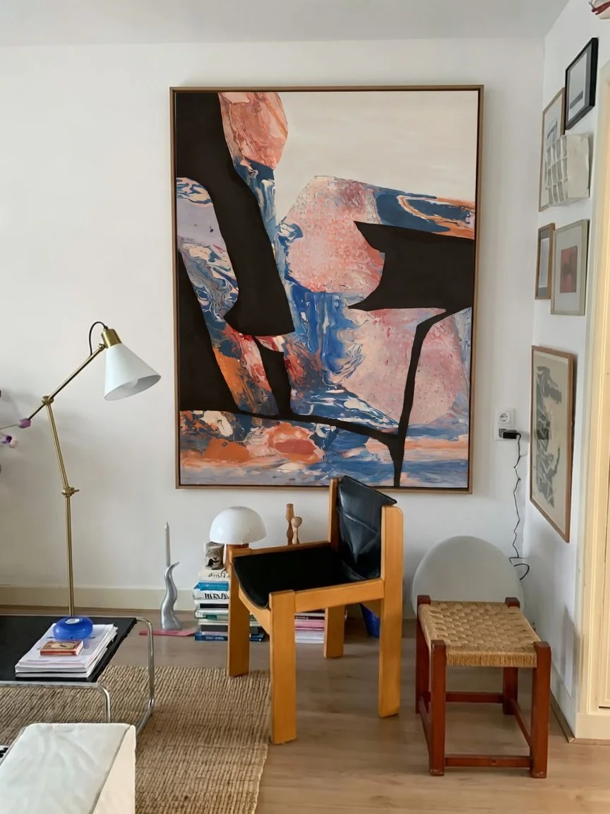 A modern living room corner featuring a large abstract painting with black, blue, pink, and white colors, a wooden chair with black cushions, a small stacked table with books and a white lamp, a floor lamp, a small woven stool, and framed art on the 