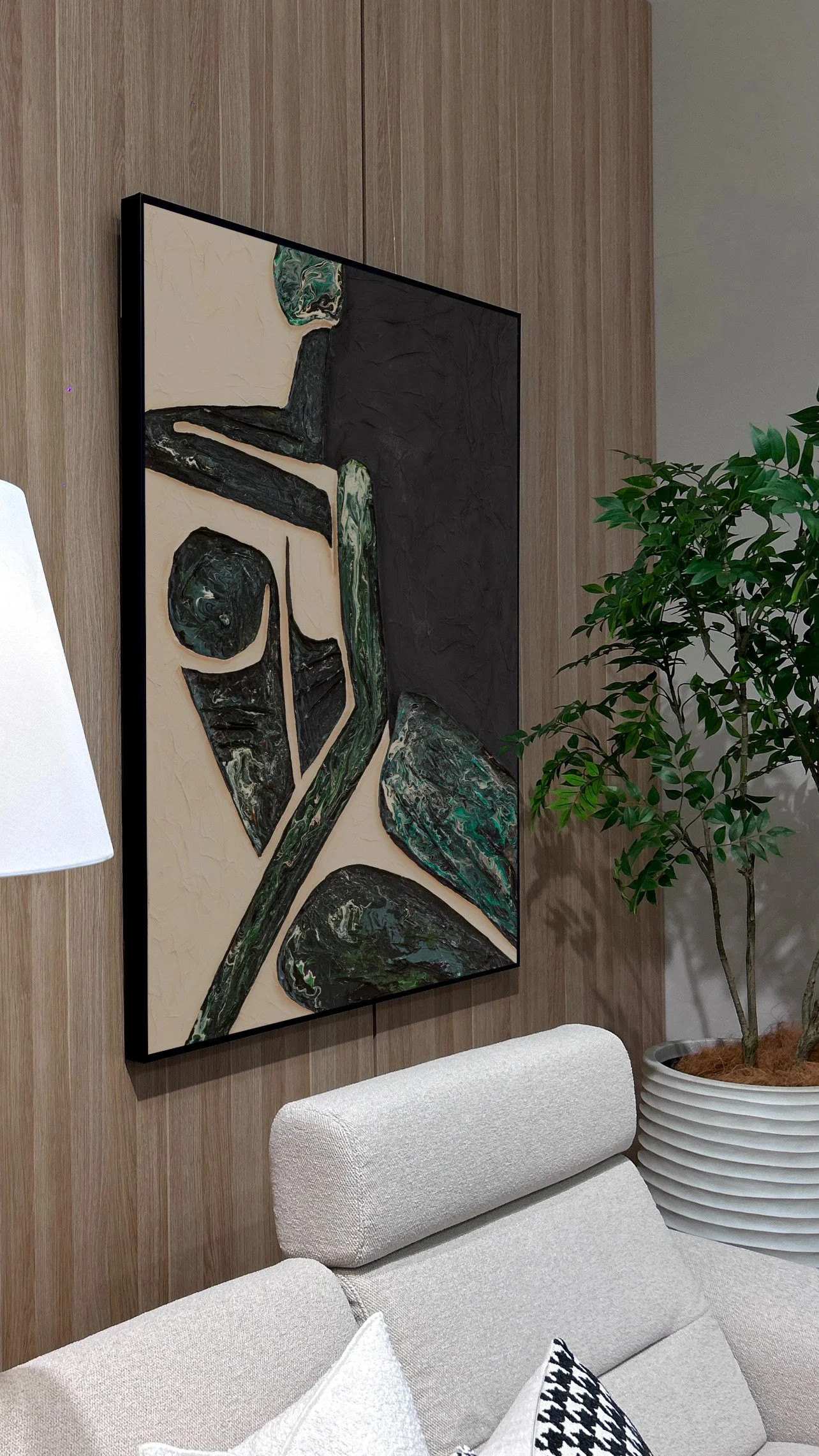 A modern living room corner features a framed abstract painting with black, green, and cream colors hanging on a wooden-paneled wall. To the right, there's a potted green plant with glossy leaves in a ridged white pot. A white upholstered sofa with p