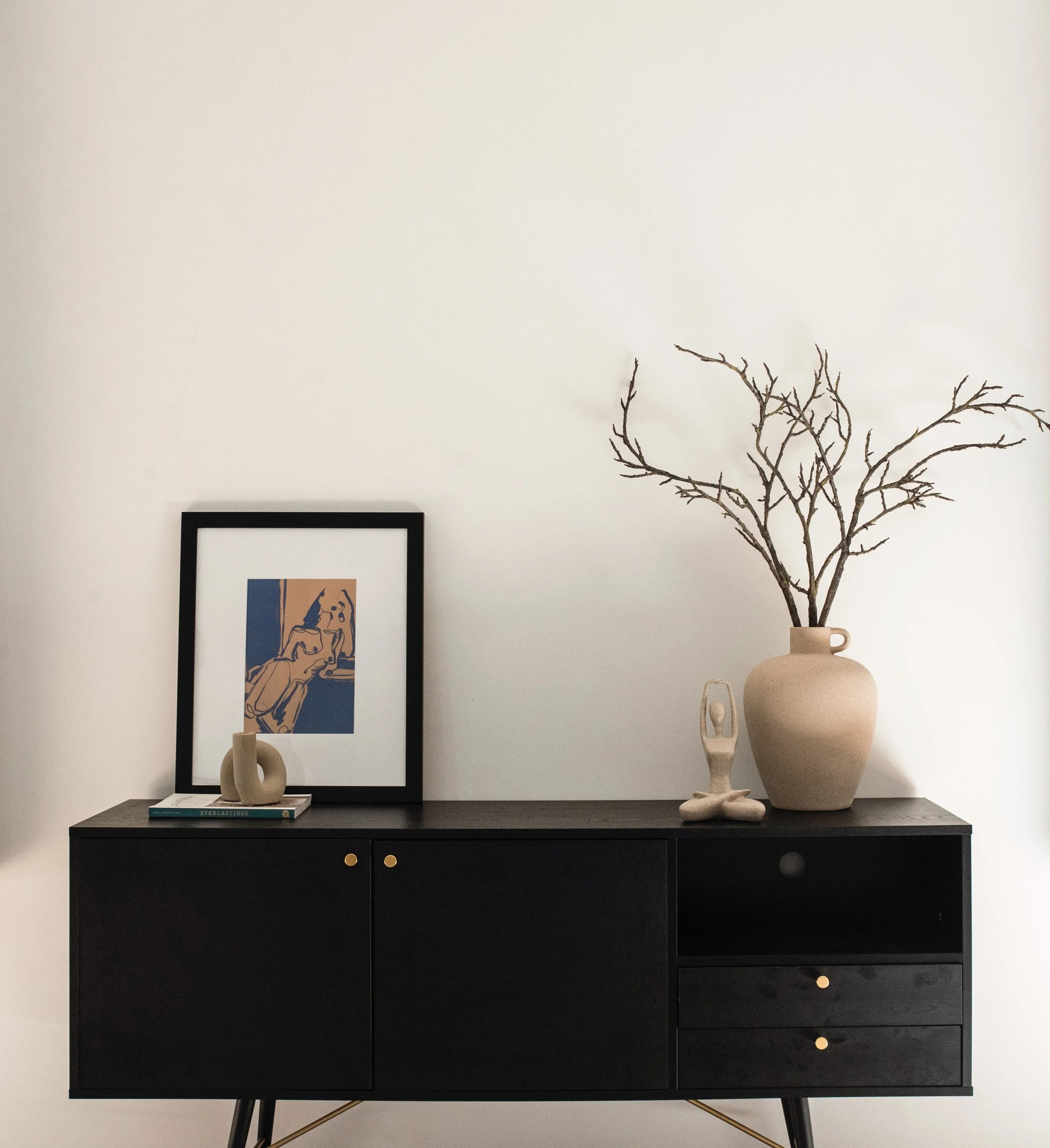 Black sideboard with three drawers and two cabinets, decorated with a large beige vase with bare branches, an abstract framed artwork, and a small beige sculpture, against a white wall.