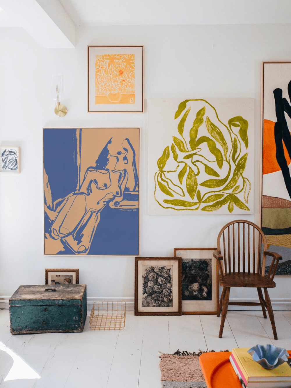 Interior of an art gallery with various framed artworks on a white wall, including illustrations of a hand holding a cigarette, a plant with yellow leaves, and abstract shapes. There is a wooden chair, an old painted chest, smaller unframed artworks 